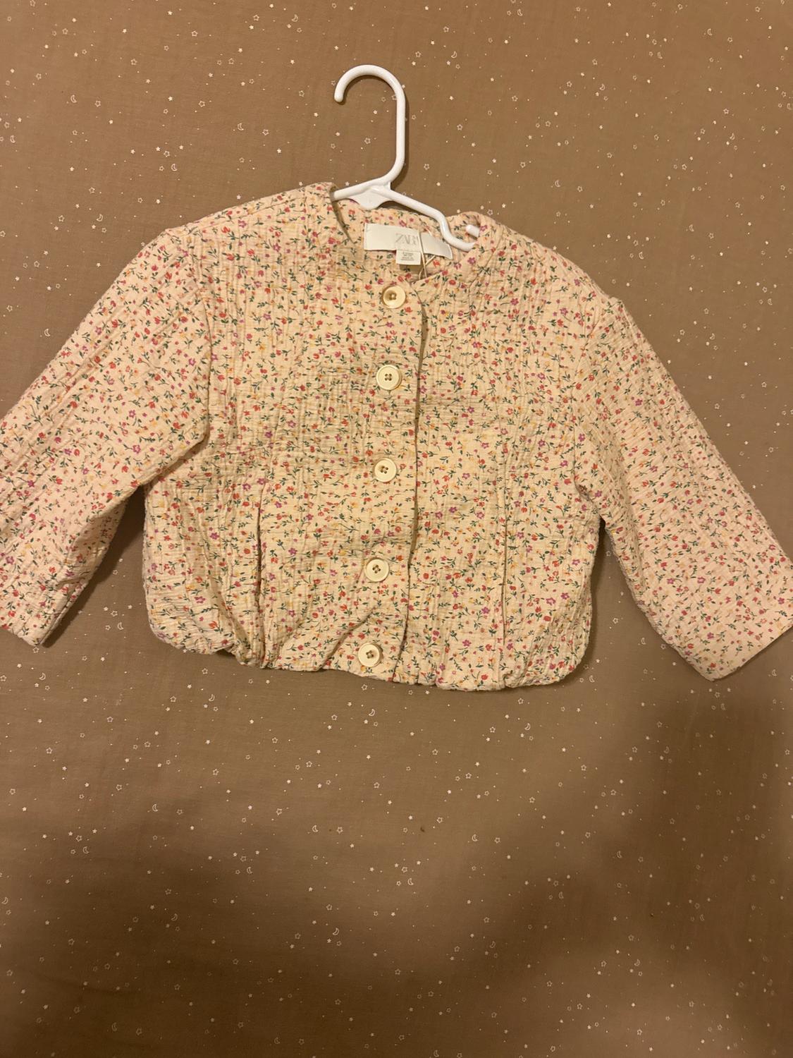 Child's jacket made from patterned fabric with small flowers, large buttons, and a beige background. Hung on a white hanger against a brown background.