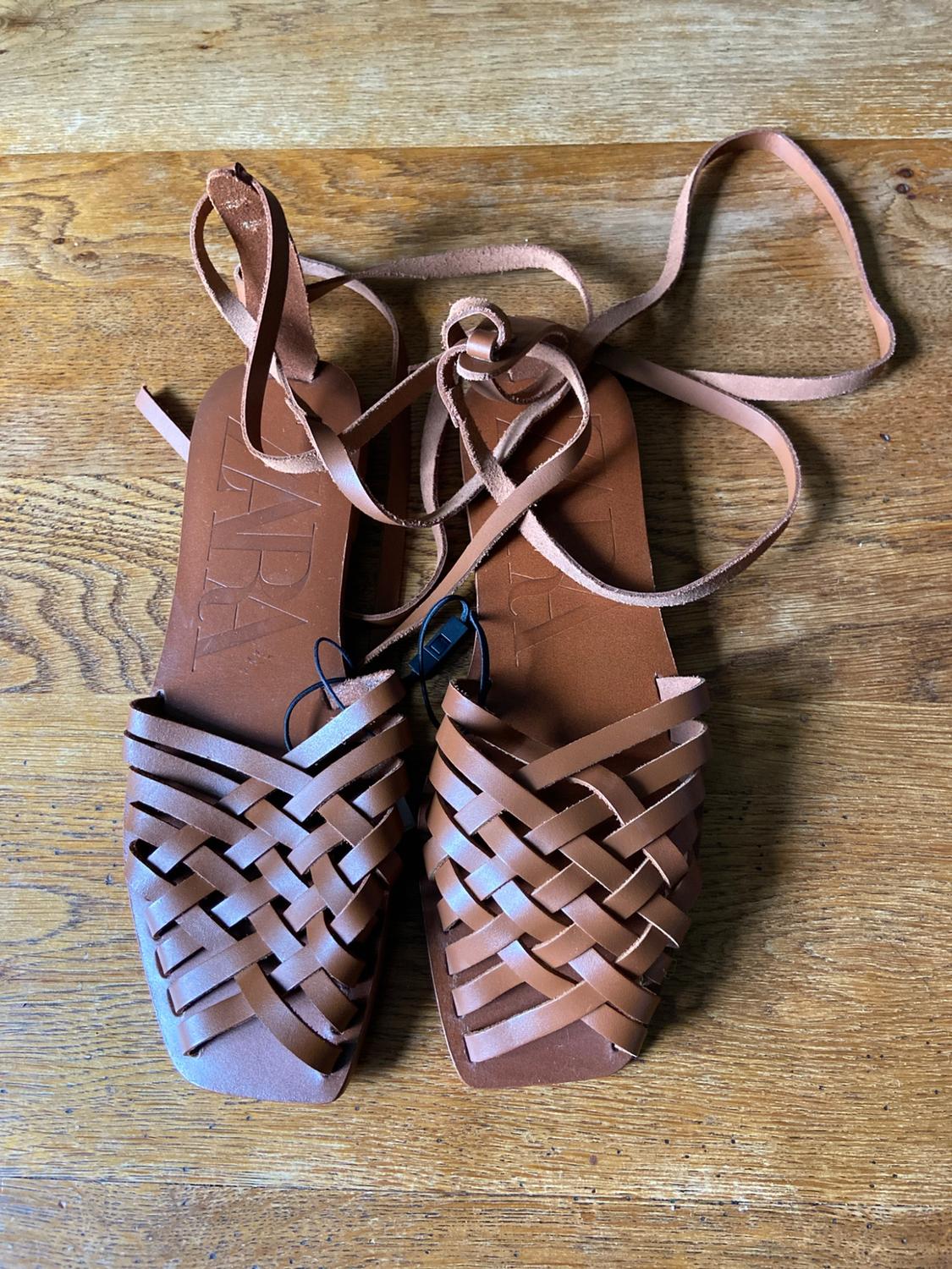 Brown flat sandals with a braided design and lace-up straps, on a wooden surface. ZARA text on the insole.