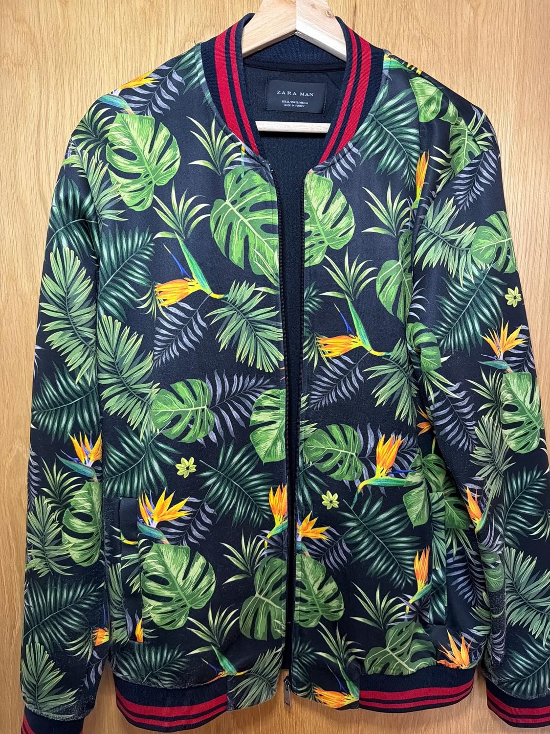 Jacket with a tropical print of green leaves and orange flowers on a dark background, hanging on a wooden hanger.