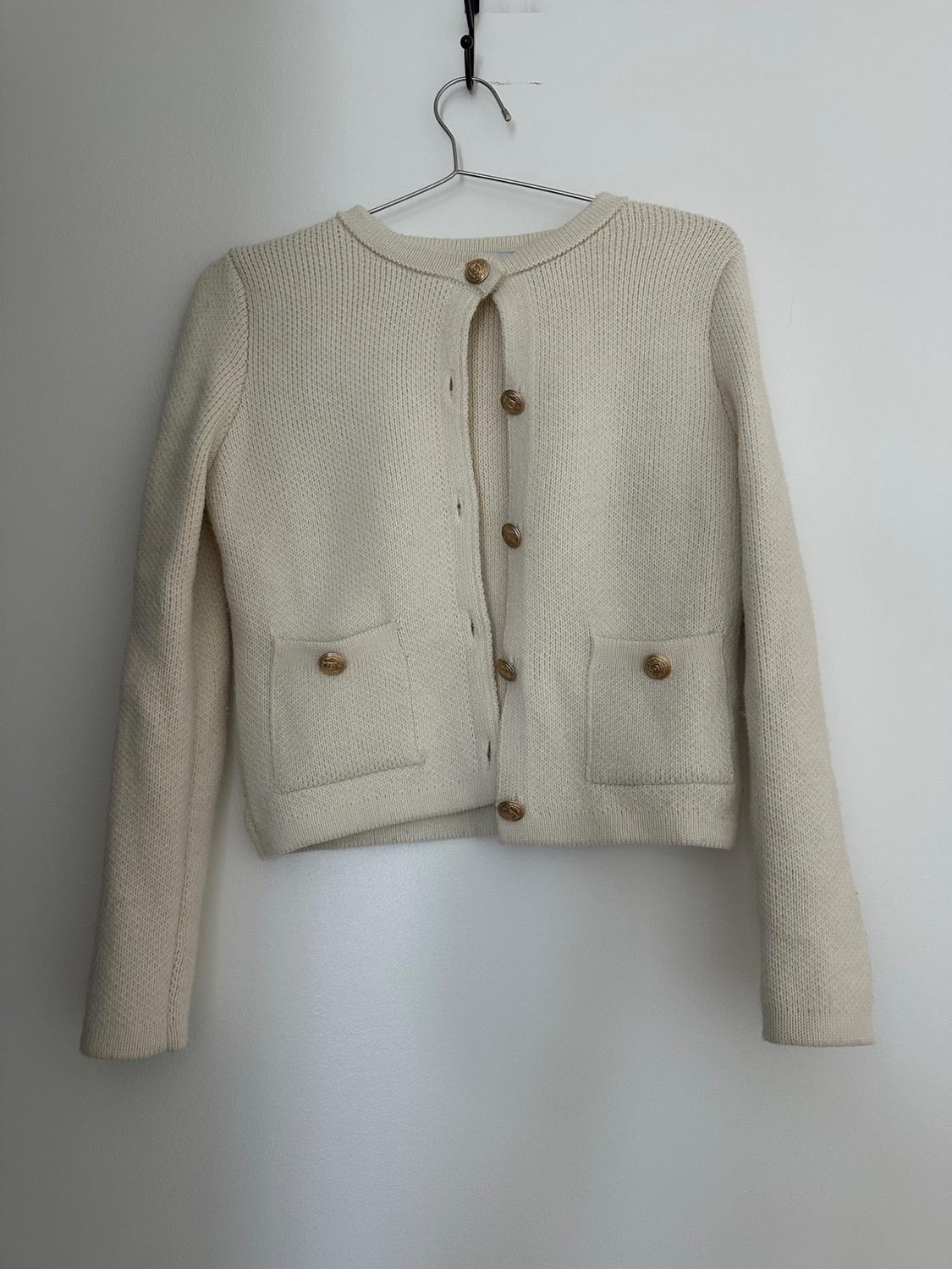Cream knit cardigan with gold buttons and front pockets, hanging on a wire hanger against a white background.