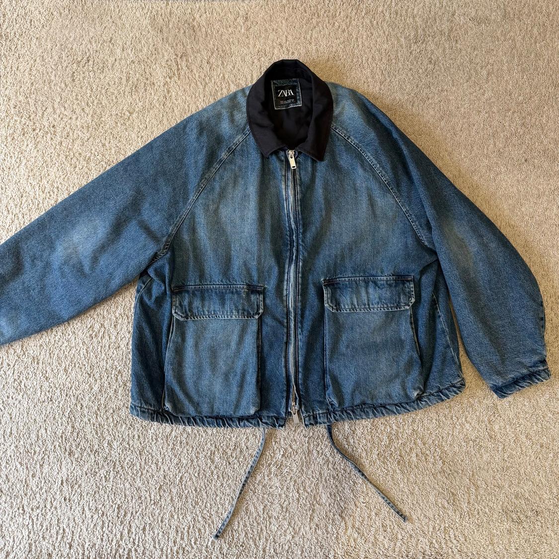 Blue denim jacket with a front zip and two large pockets, on a beige carpet. Visible label: "ZARA".
