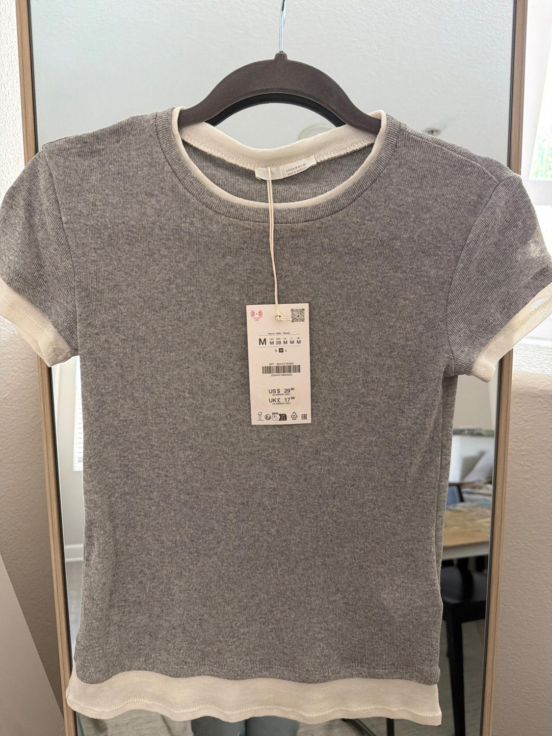 Grey short-sleeved T-shirt with white trim on a black hanger, with a price tag and size M, reflected in a mirror.