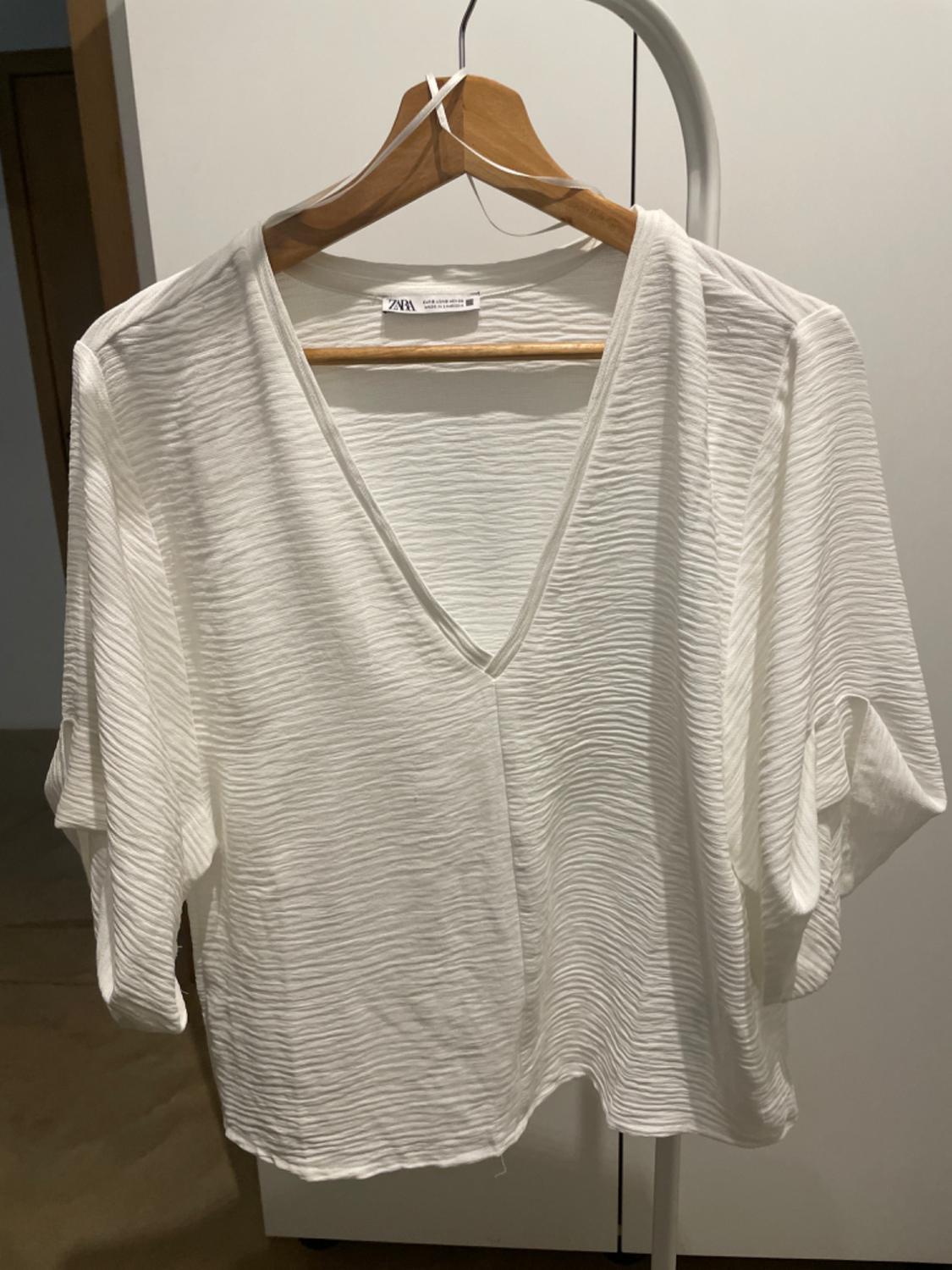 White short-sleeved blouse with crinkled texture, hanging on a wooden hanger. Visible label with text "Zara".