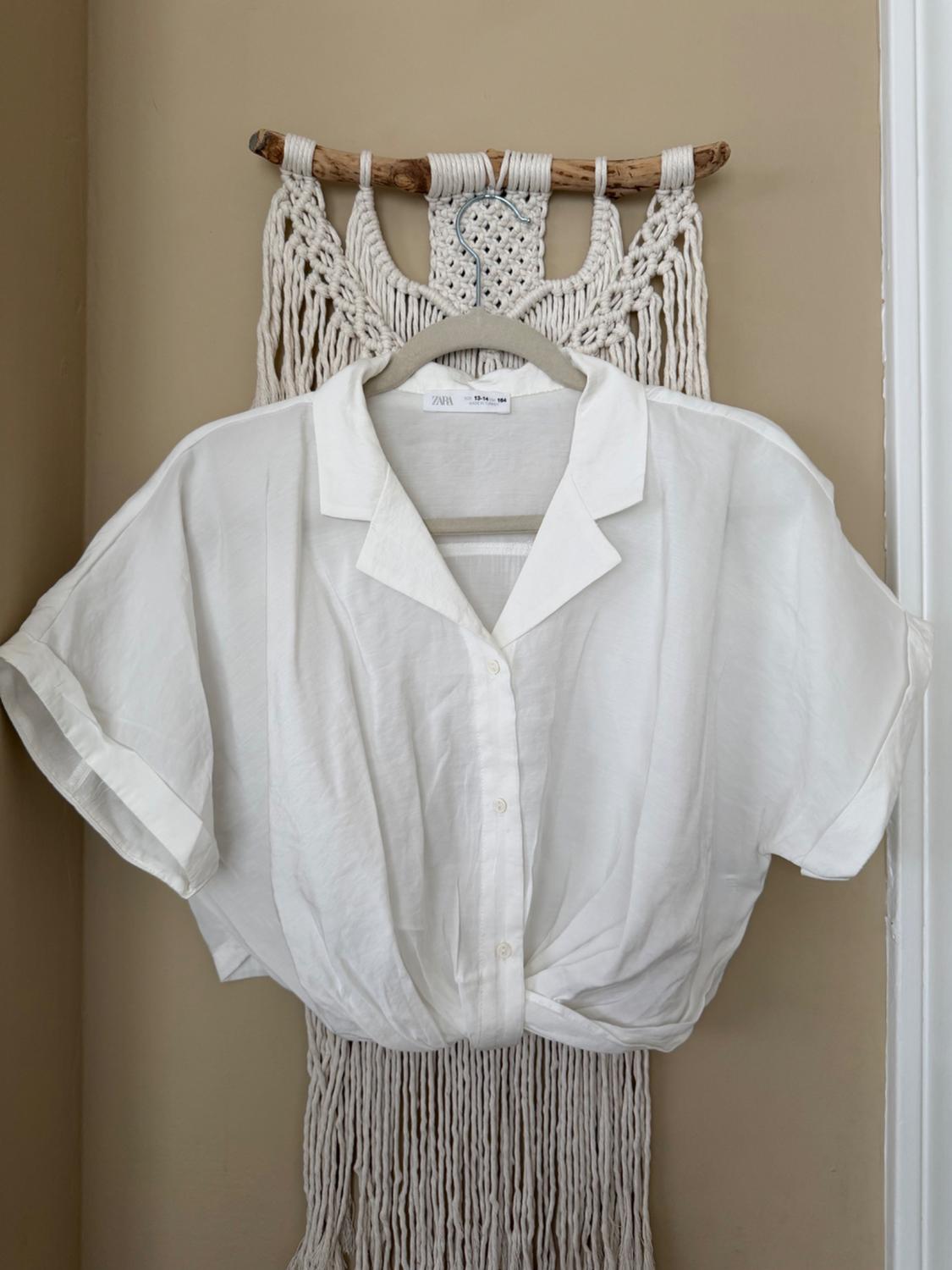 Short-sleeved white shirt hanging on a macrame hanger against a beige wall. Visible tag: Zara.