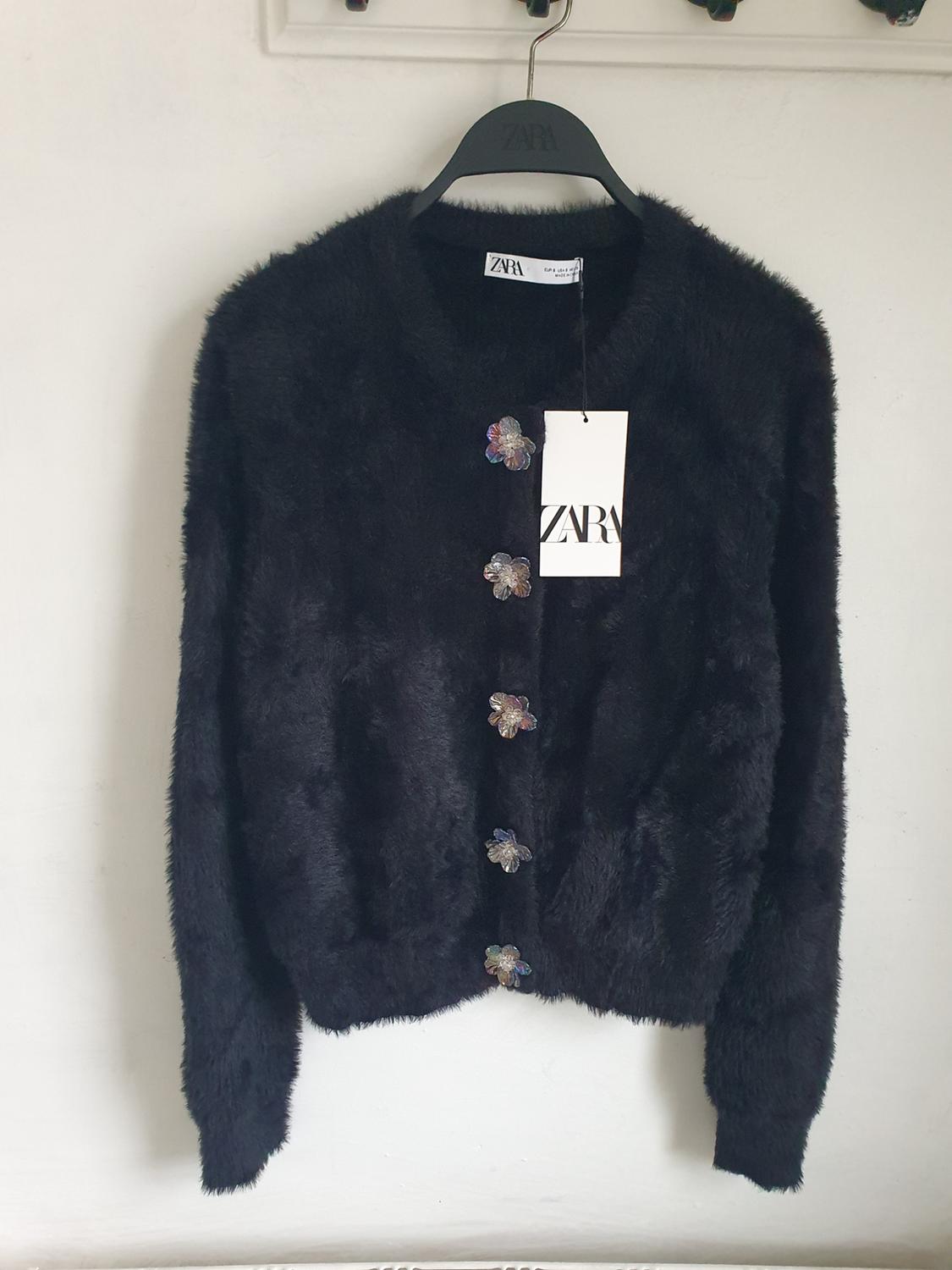 Black faux fur jacket with shiny floral buttons, hanging on a black hanger with a visible Zara tag.