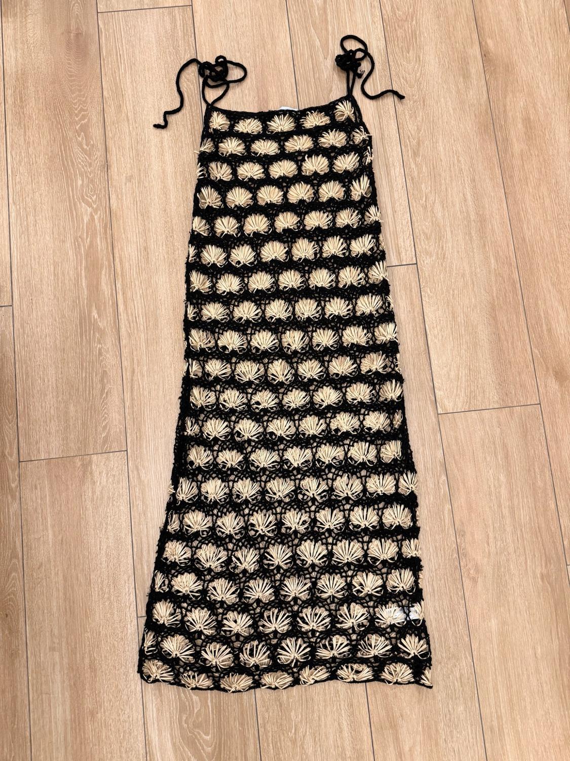 Black open-knit maxi dress with beige fan patterns and slim tie-up straps, on a light wood floor.