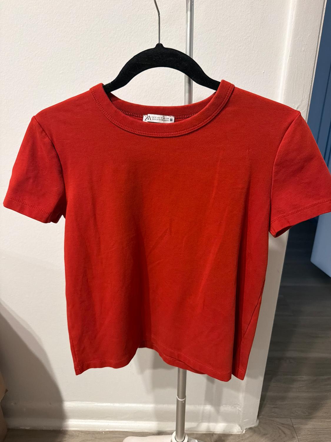 Basic red short-sleeved crew-neck t-shirt, hanging on a black hanger on a metal clothing rack in front of a white wall.