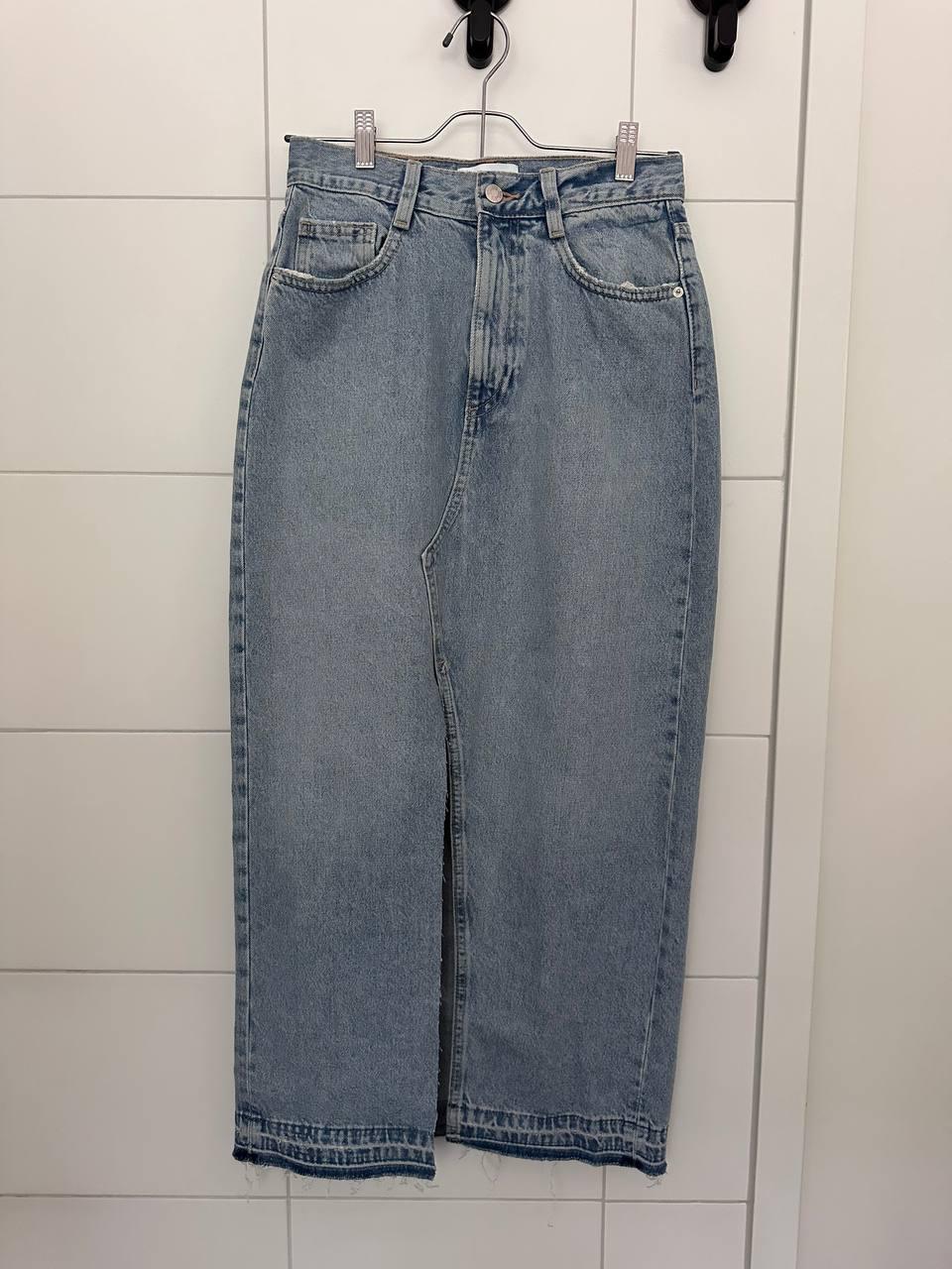 Light blue long denim skirt with a front slit and frayed hem, hanging on a hanger against a white tiled wall.