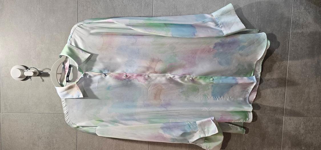 Long-sleeved shirt with a watercolour print in pastel tones, hanging on a hanger against a background of grey tiles.