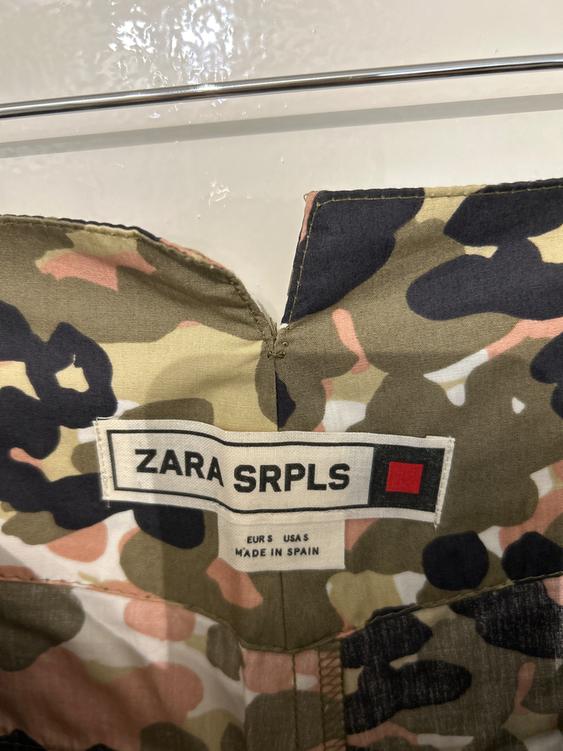 Clothing label with camouflage print in green, black, and pink tones. Text: "ZARA SRPLS, EUR S USA S, MADE IN SPAIN".