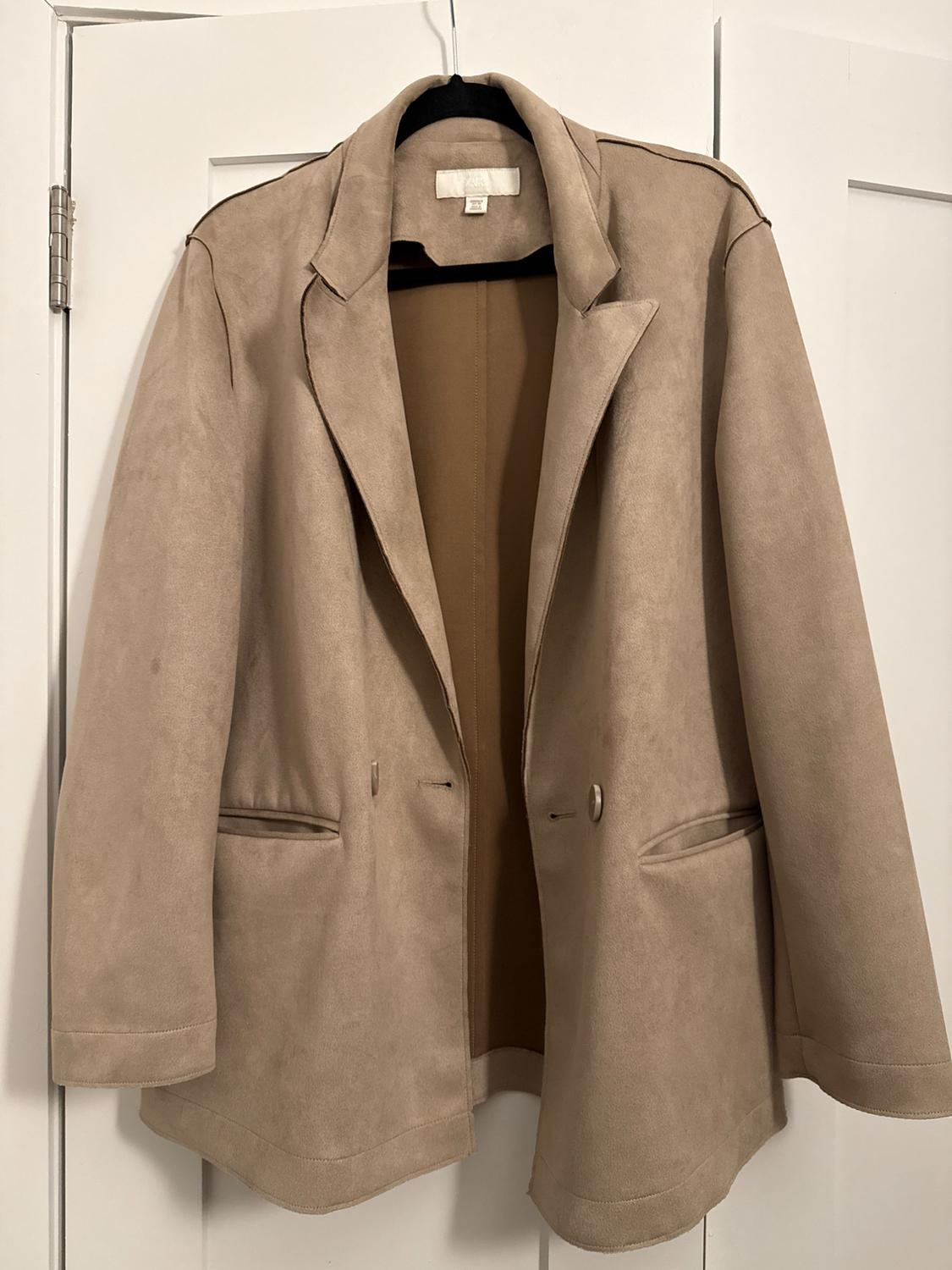 Beige jacket hanging on a black hanger over a white door. It has lapels, two pockets, and front buttons.