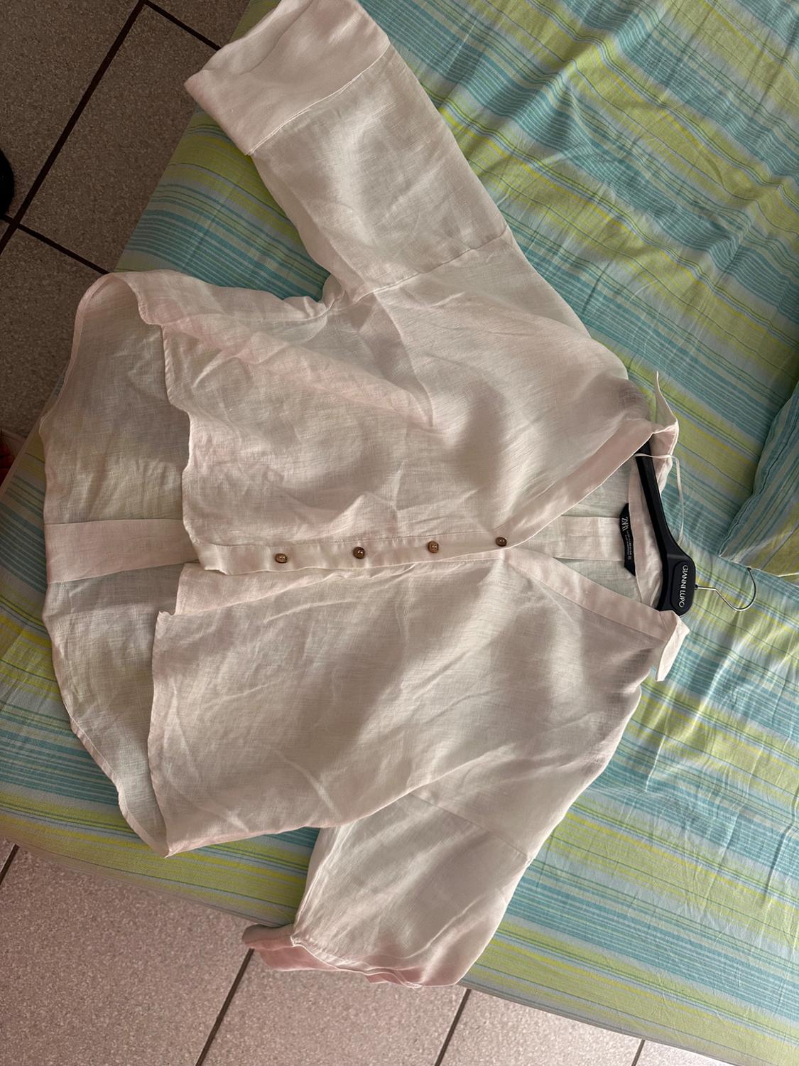 White button-down shirt on a bed with striped sheets. Hanger with GIANNI LUPO text and ZARA label. Relaxed atmosphere.