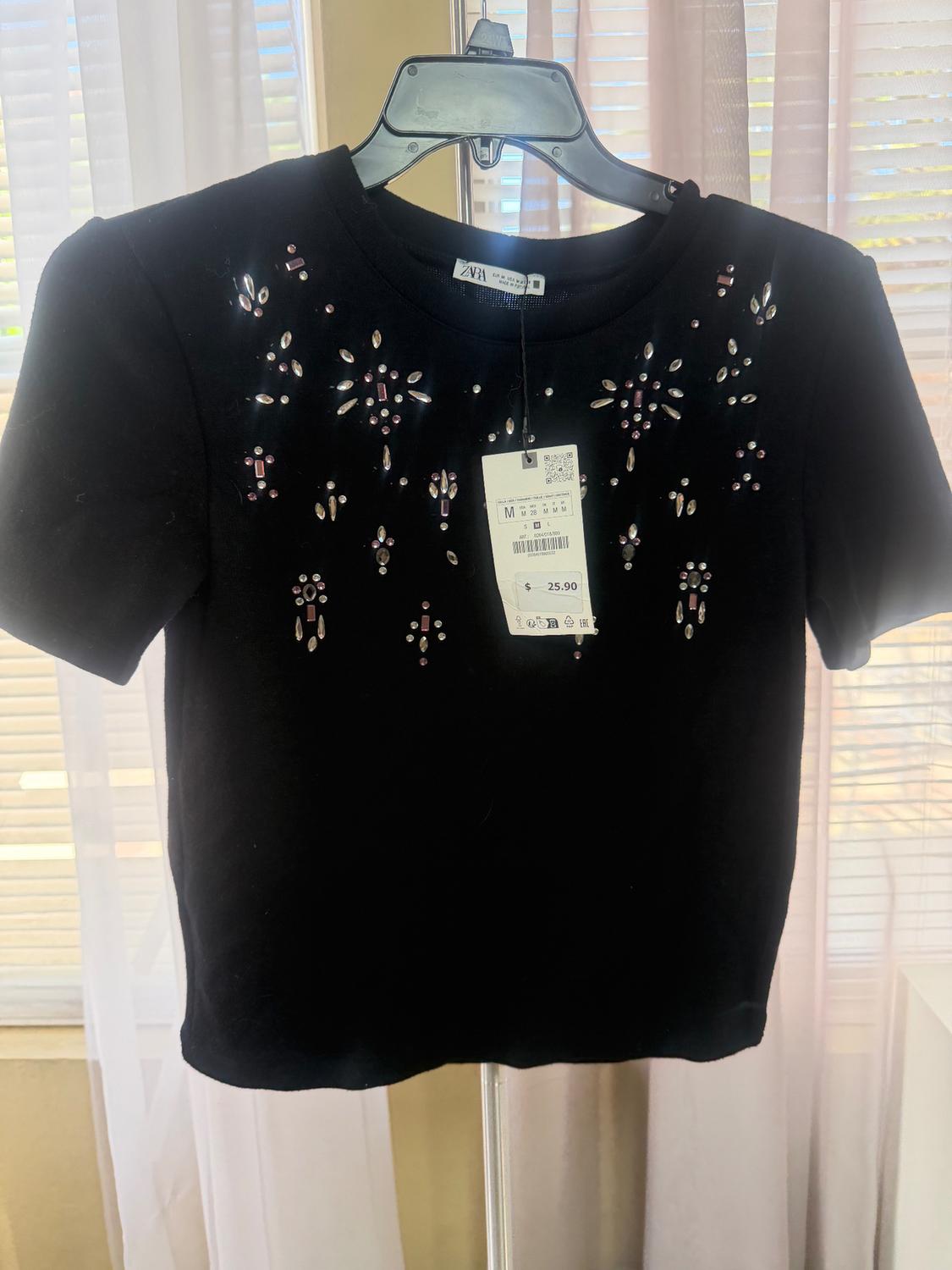 Black t-shirt with sparkly embellishments on the chest, hanging on a hanger. Visible tag with a price of 25.90. Curtains in the background.