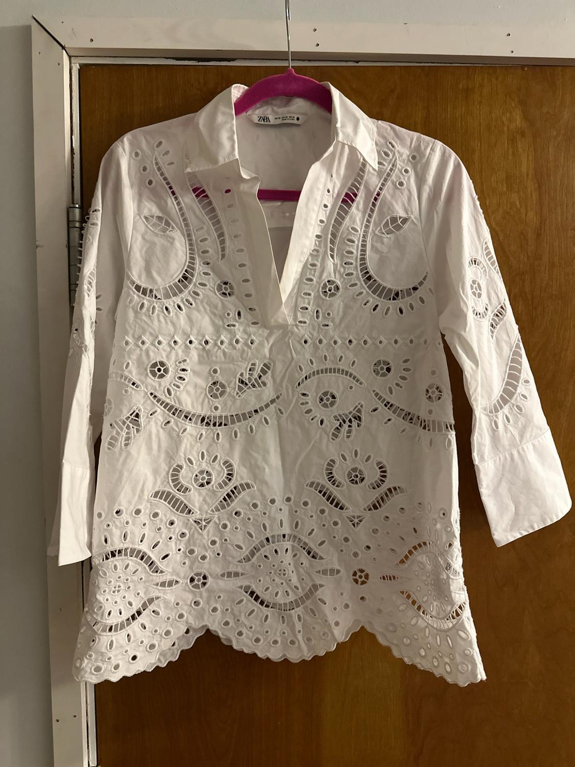 White blouse with cut-work embroidery and a V-neck, hanging on a pink hanger over a wooden door. Visible text: ZARA.