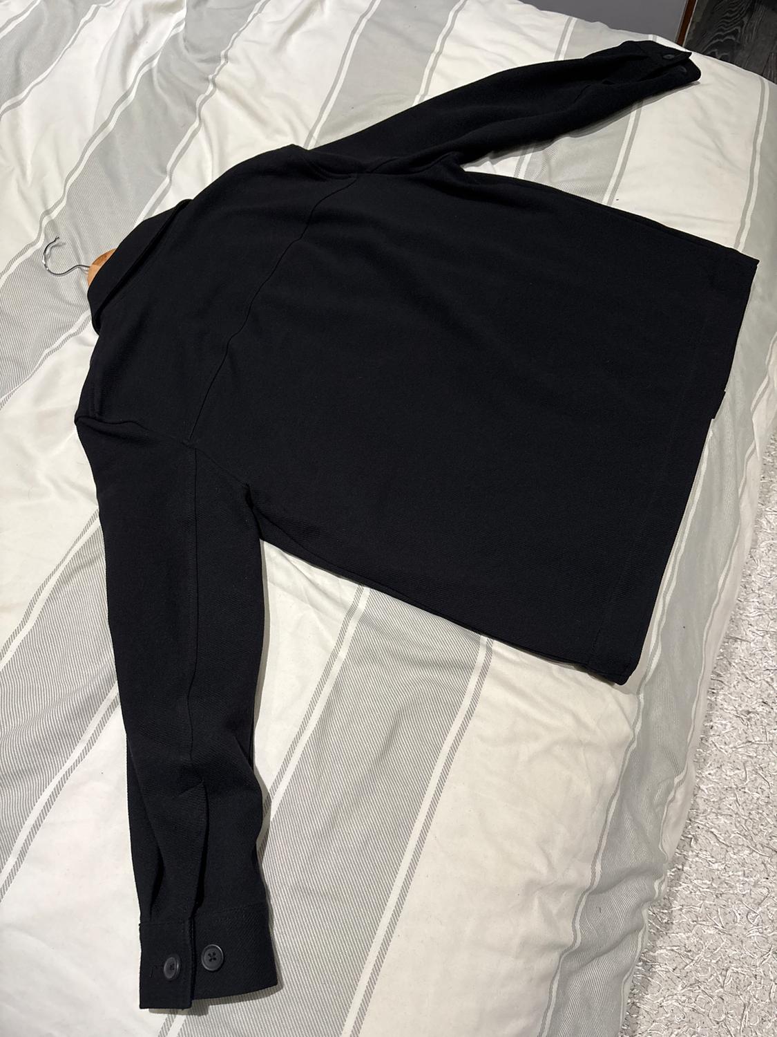 Black long-sleeved coat with buttons on the cuffs, spread out on a bed with grey and white striped sheets.
