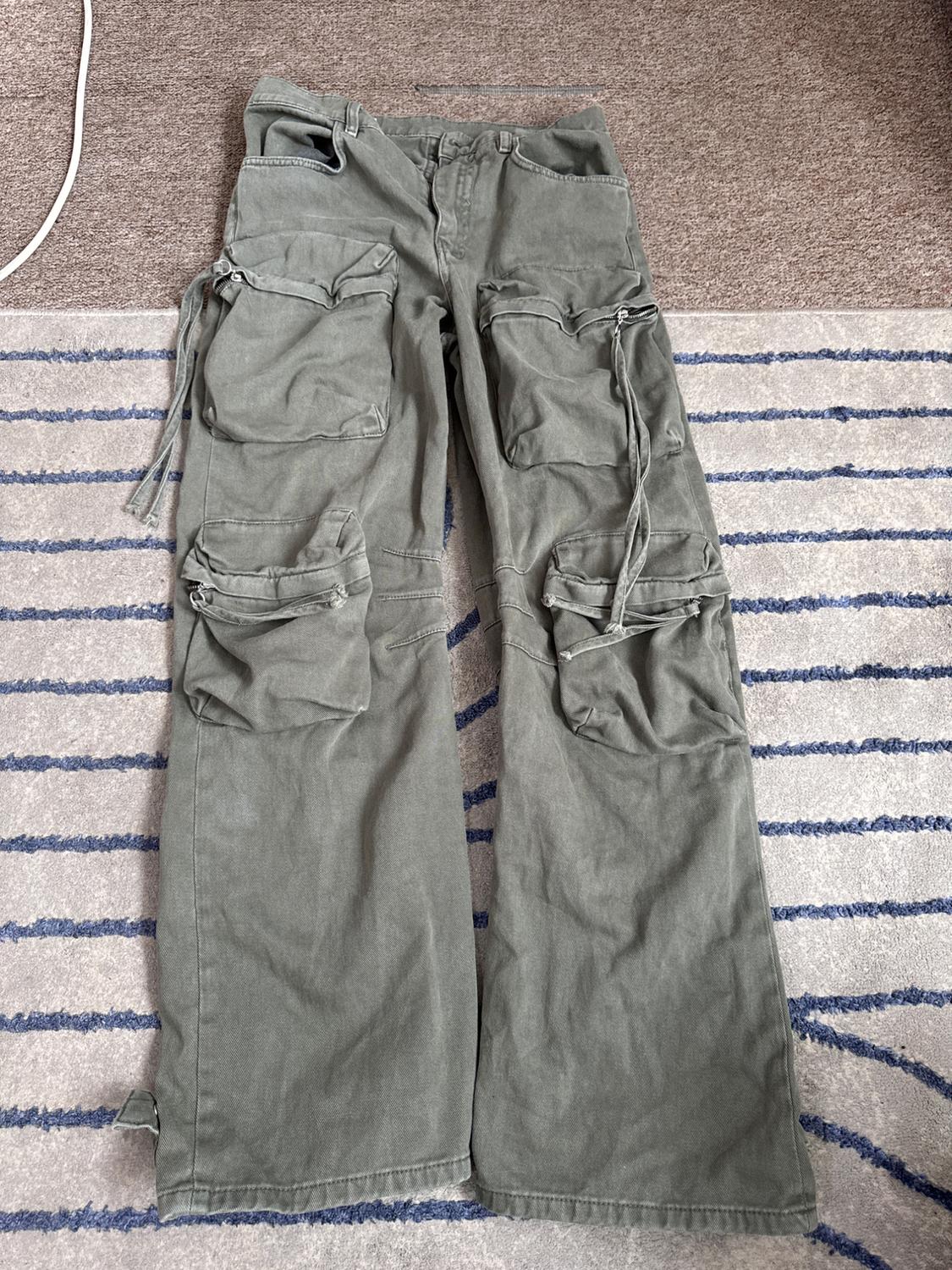 Olive green, wide-leg cargo trousers with large front pockets and straps, on a blue striped rug.
