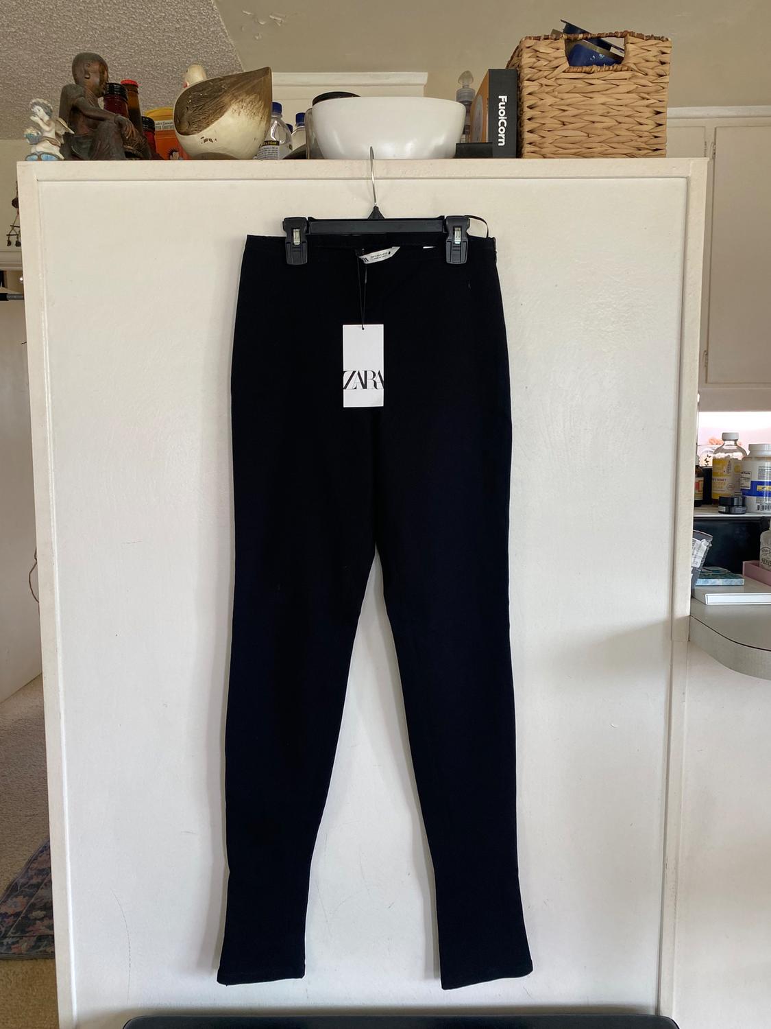 Black pants hanging on a hanger with a Zara tag, against a kitchen background with decorative objects and boxes.