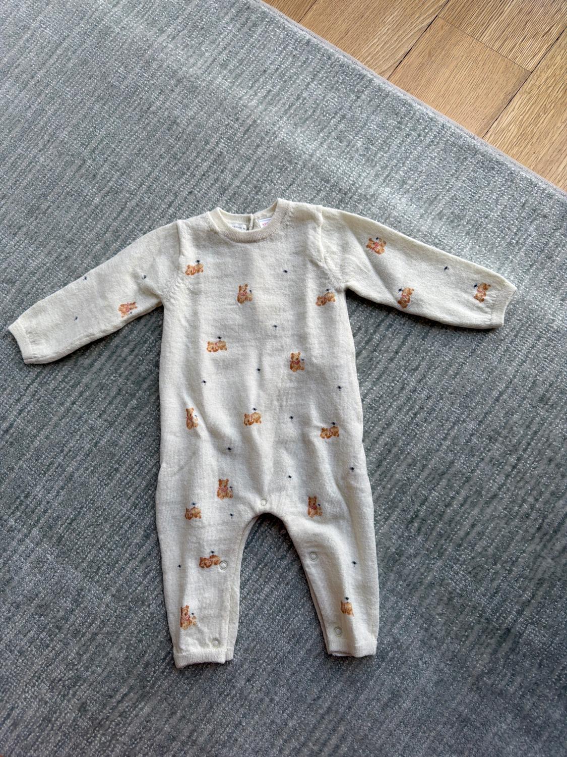 Beige babygrow with brown bear print on a grey rug. Long sleeves and poppers at the crotch.