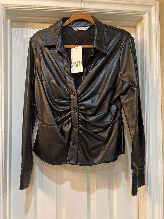 Black faux leather shirt with front ruching, hanging on a wooden hanger. Visible tag with the text "ZARA".
