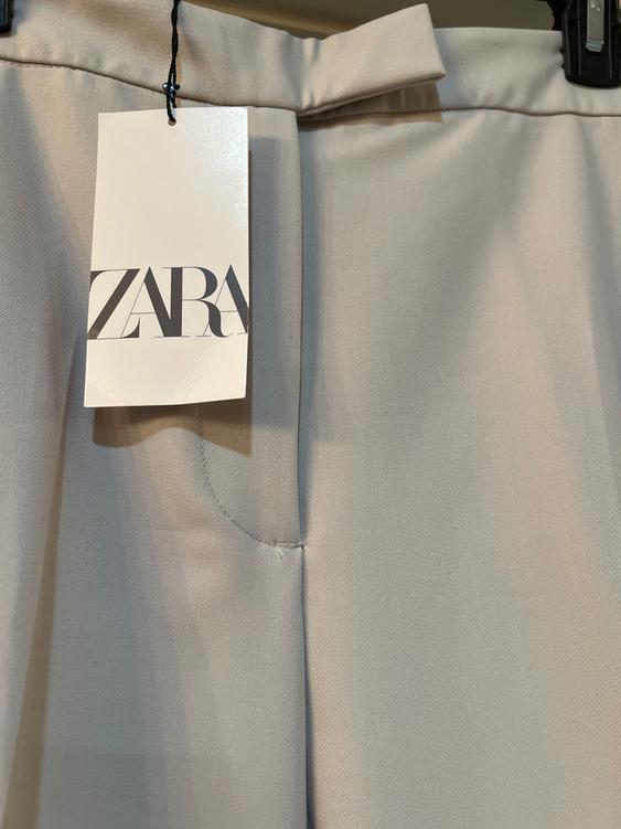 Zara tag hanging from beige pants on a hanger. The tag is white with the Zara logo in black letters.