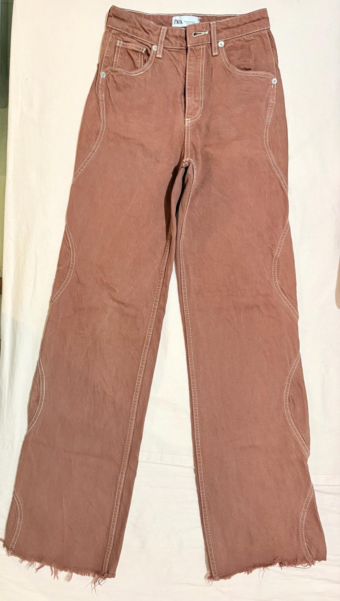 Brown trousers on a light background with white stitching, curved leg design and frayed hem. ZARA label.