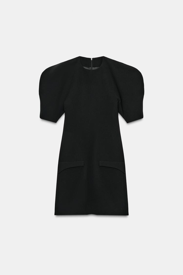 Front view of a short black dress with a round neck, short voluminous sleeves, front pockets, and a back zip fastening.
