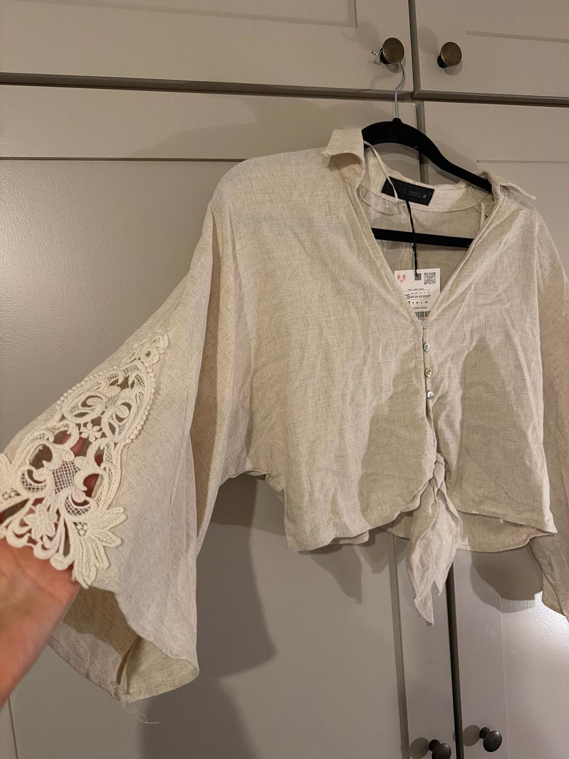 Beige blouse with buttons and a knot at the front, hanging in a wardrobe. A hand stretches out the wide sleeve to display its lace trim.