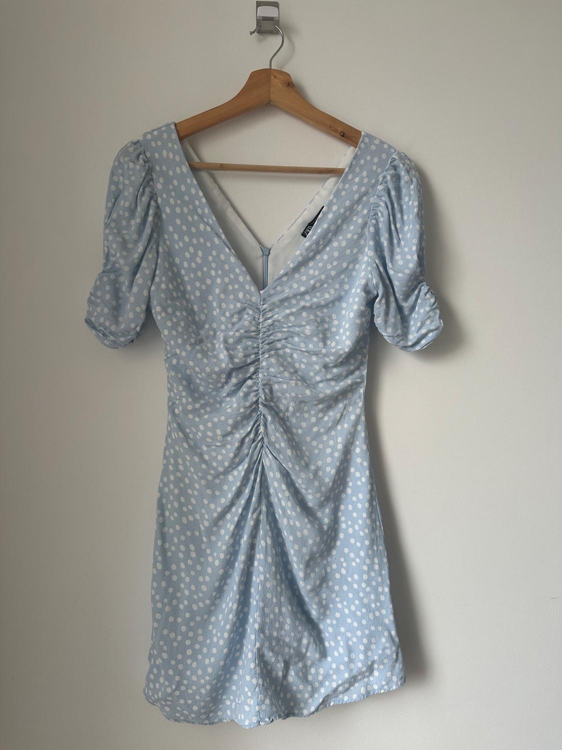 Light blue polka dot dress with white spots, V-neckline, short puffed sleeves and front ruching, hanging on a wooden hanger.