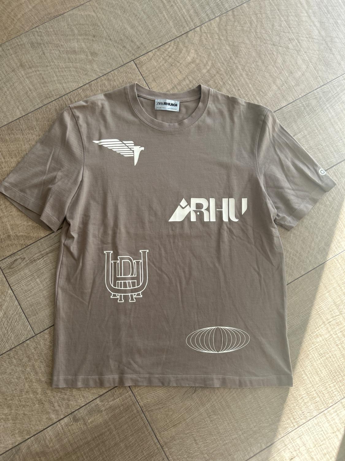 Brown T-shirt with white graphics of wings, "ARHU", a symbol, and an oval design. "ZNR HUGI" tag on the collar. Light wood background.