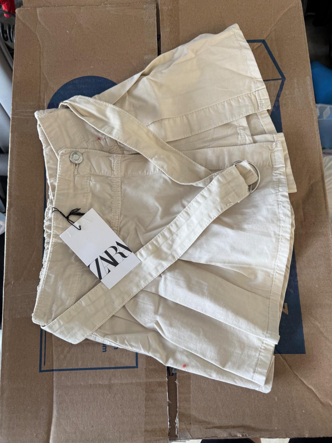 Detail of beige shorts with belt on a cardboard box, visible label with text 'ZARA'.