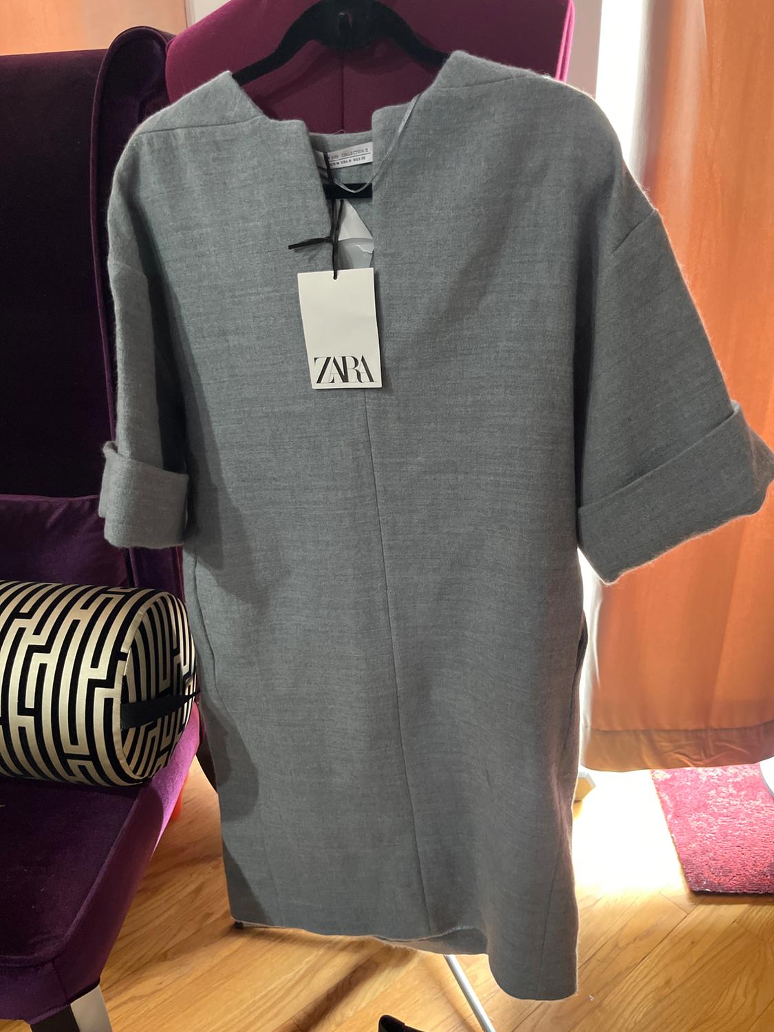Grey short-sleeved dress hanging on a black hanger with a Zara tag. Background with a purple chair and a patterned cushion.