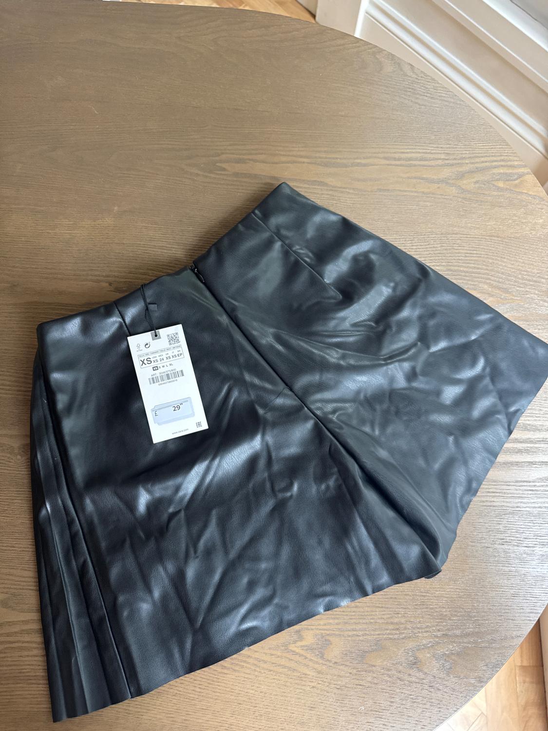 Black skirt made of synthetic material on a wooden table, with a visible label indicating size XS and a price of 29 euros.