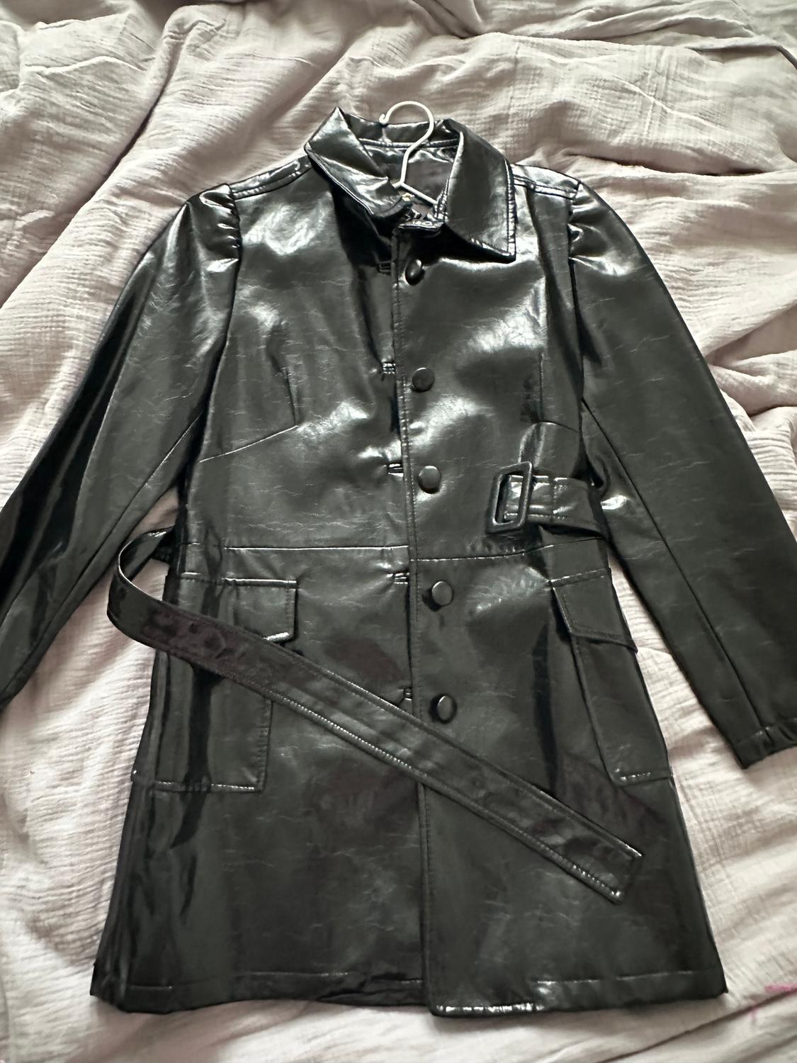 Shiny black coat with a belt and buttons, hung over a bed with a light bedspread.