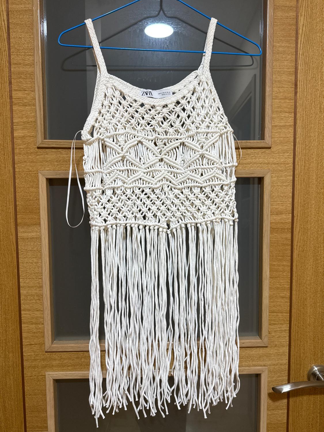 White macrame top with fringes hanging on a blue hanger in front of a wooden door with glass panels. Visible tag: Zara.