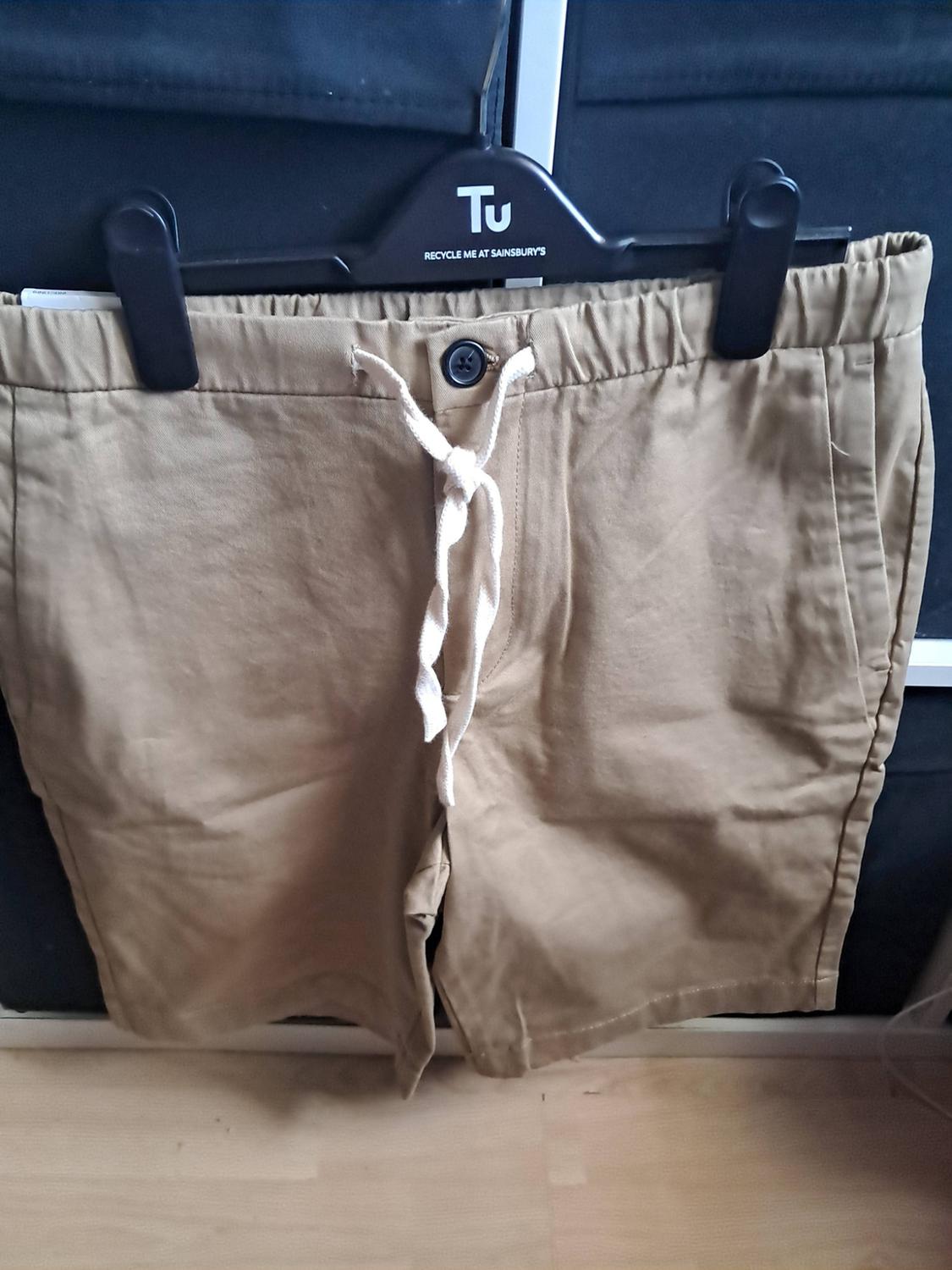 Beige shorts with a white drawstring on a black hanger. Text: Tu RECYCLE ME AT SAINSBURY'S. Casual style, dark background and wooden floor.