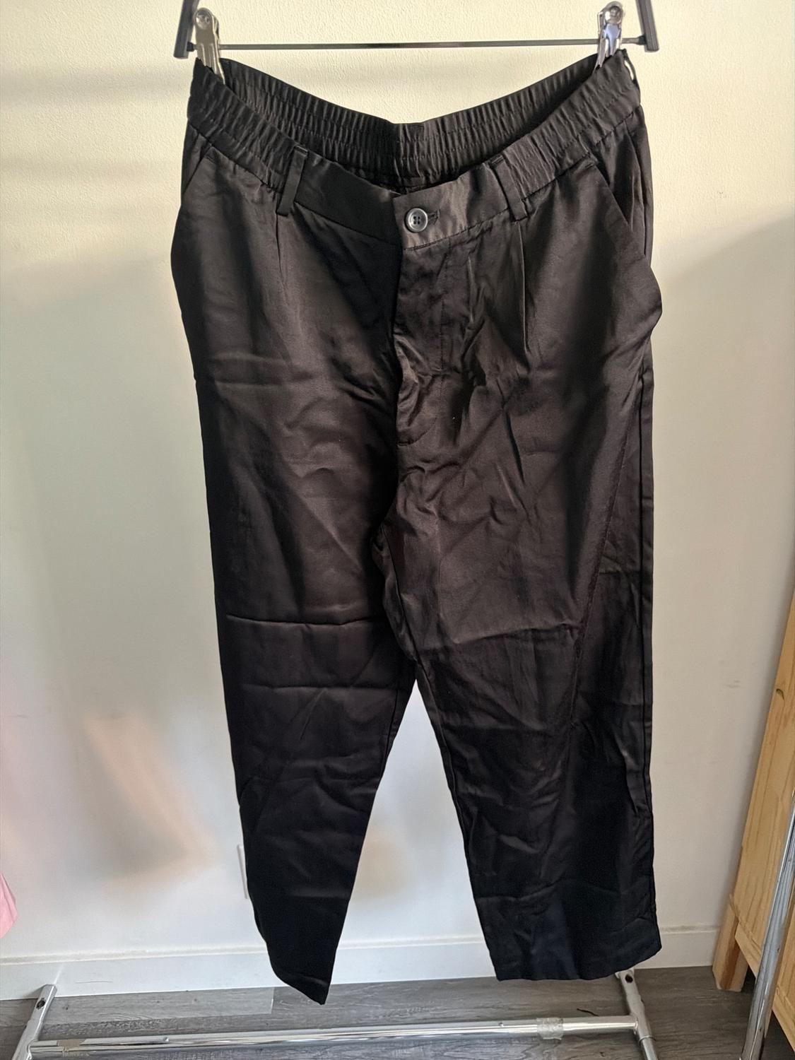Black trousers hung on a hanger, with an elasticated waist and visible button, against a light background in an indoor setting.