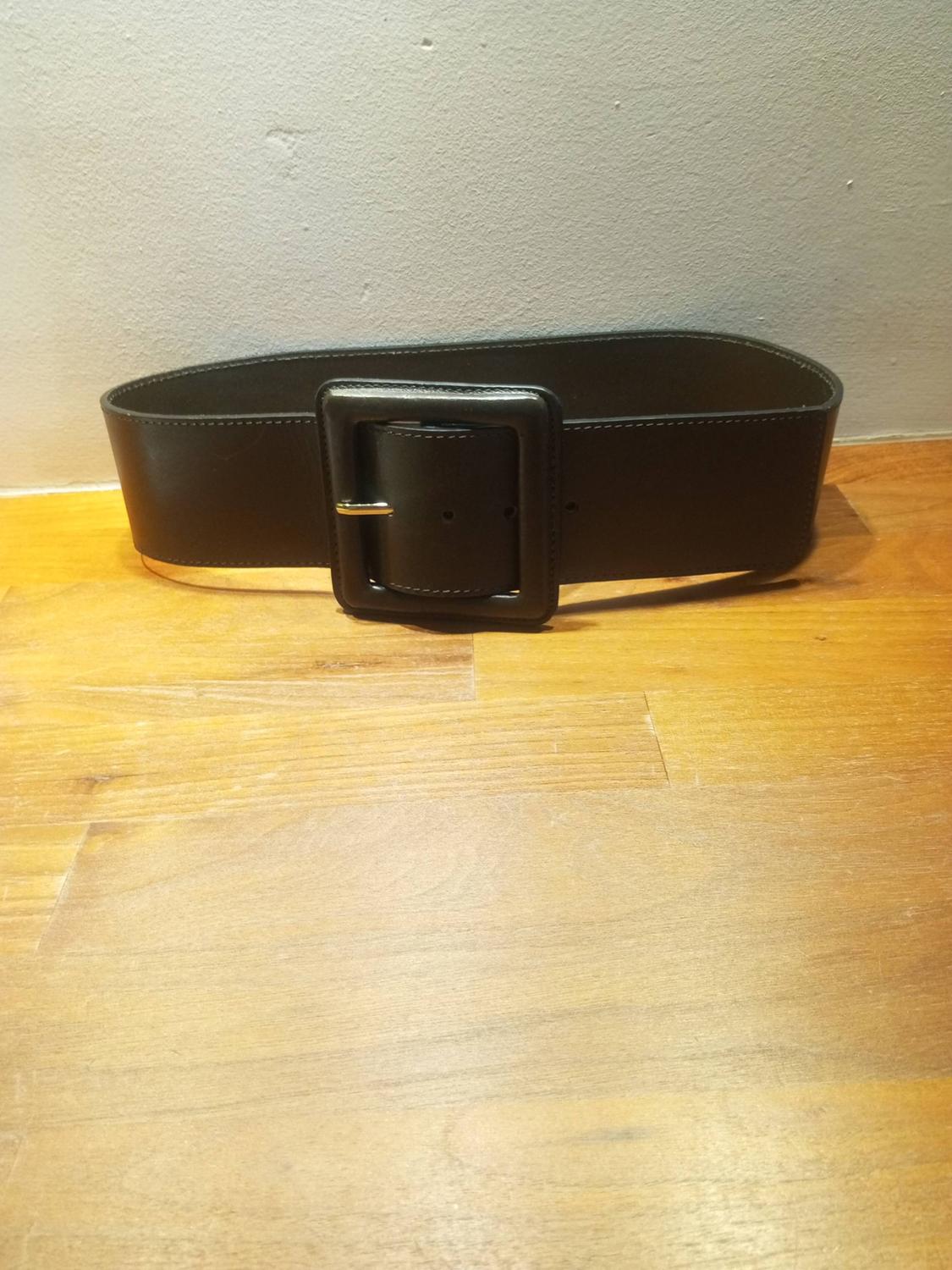 Wide black belt with a large square buckle, resting on a light wooden surface in front of a plain wall.