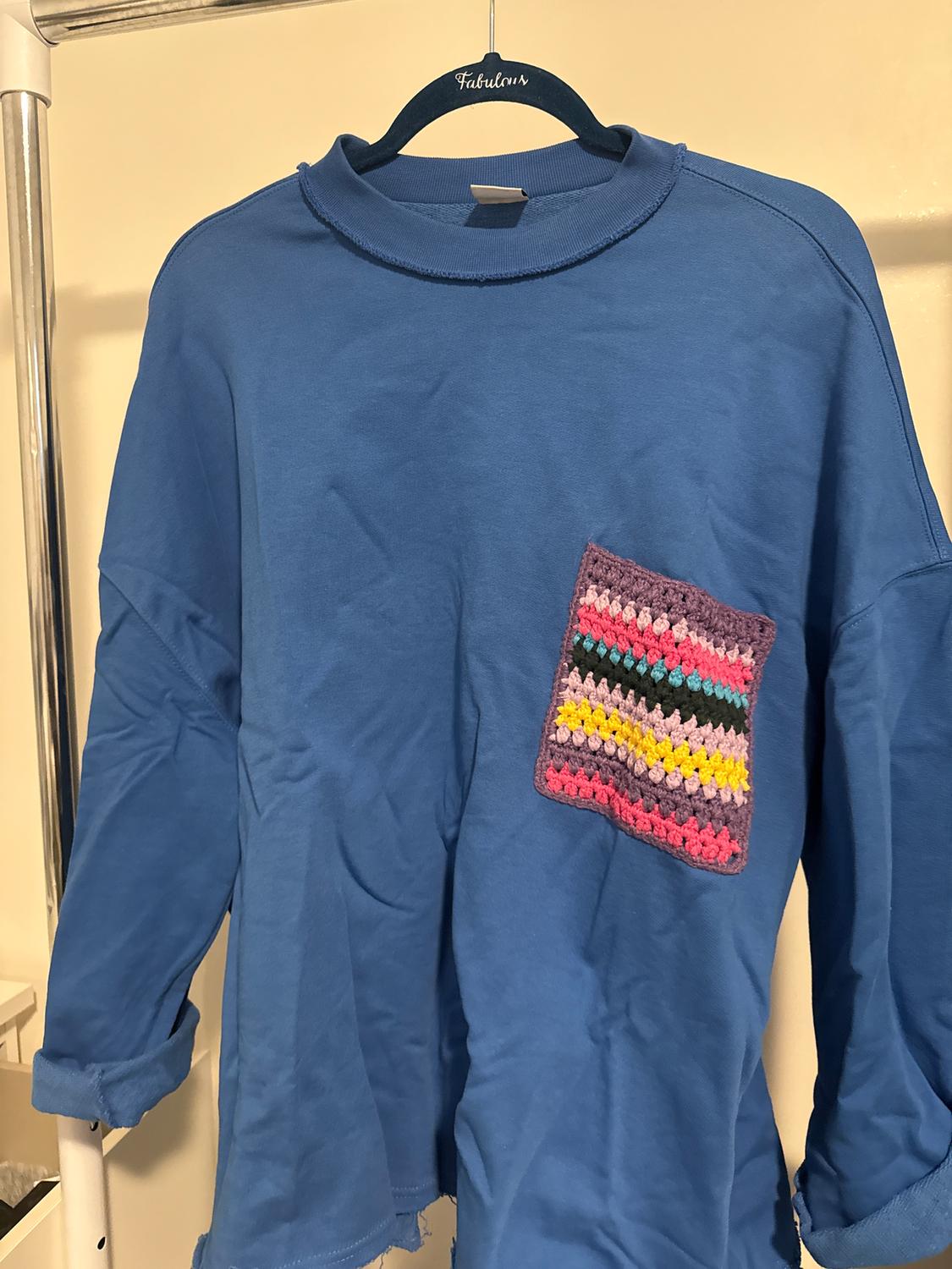 Blue t-shirt with a multi-coloured striped woven pocket, hanging on a black hanger with the word "Fabulous".