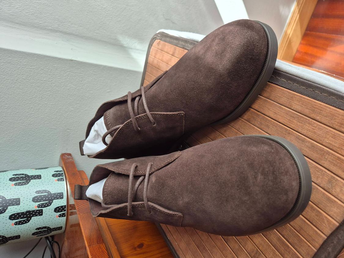 Brown suede lace-up ankle boots on a slatted wooden surface, next to a cactus-print object. Relaxed atmosphere.
