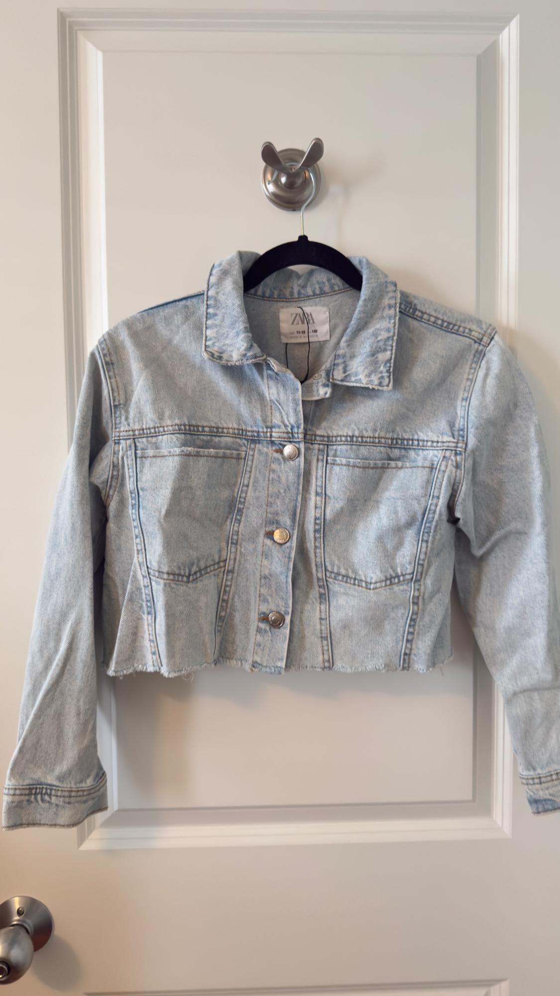 Light blue denim cropped jacket hanging on a white door. It has metallic buttons and a classic collar. Visible tag.