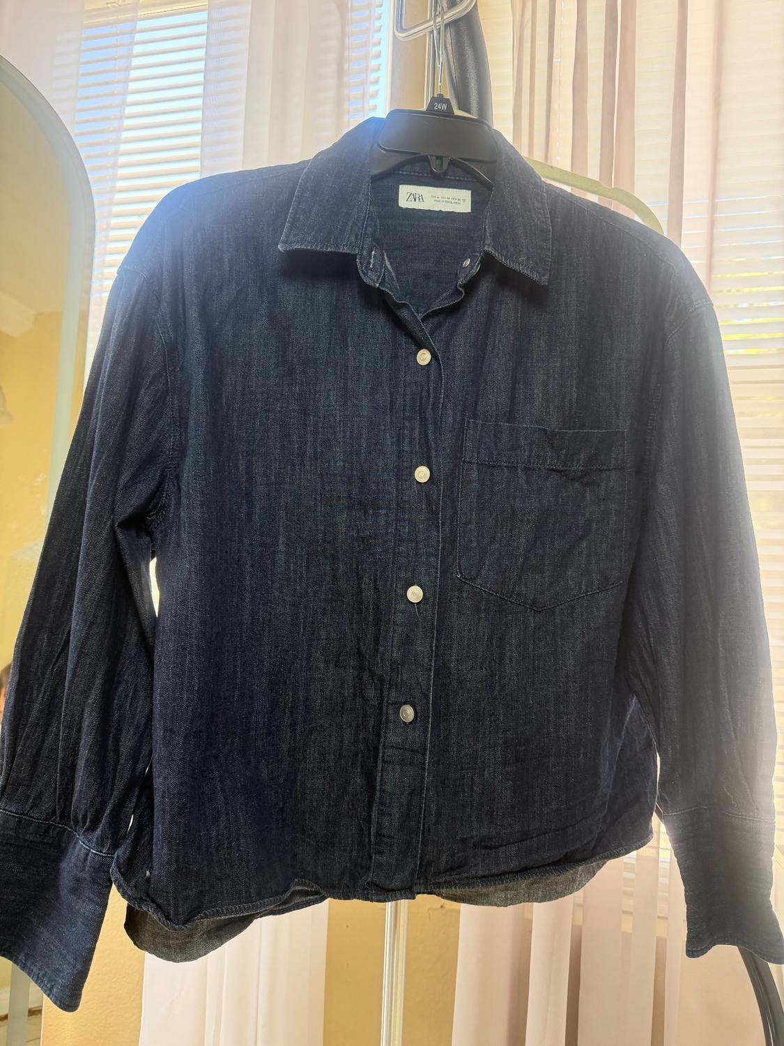 Dark blue denim shirt hanging on a black hanger, with white buttons and a chest pocket, in front of light curtains.