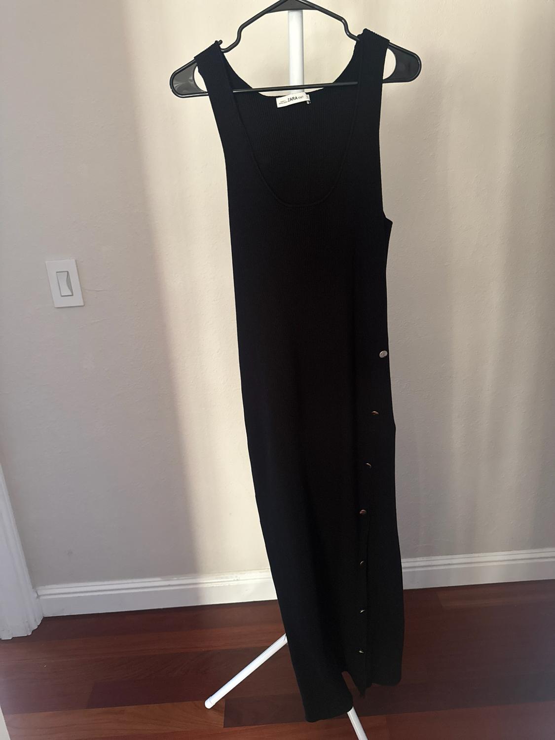 Sleeveless black knit dress with side buttons hanging on a hanger. ZARA KNIT tag. Light wall background and wooden floor.
