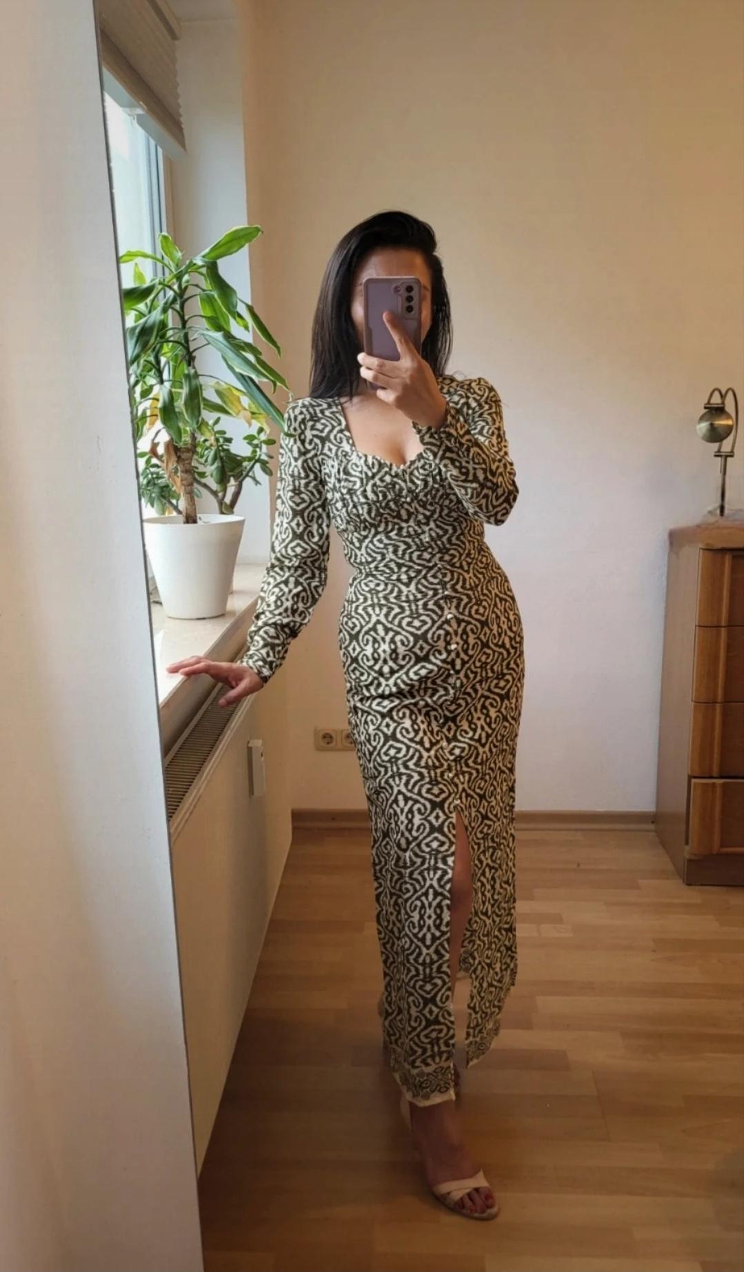 Woman wearing a long, fitted dress with a geometric print in beige and black tones, front slit, white sandals, decorative plant in the background.