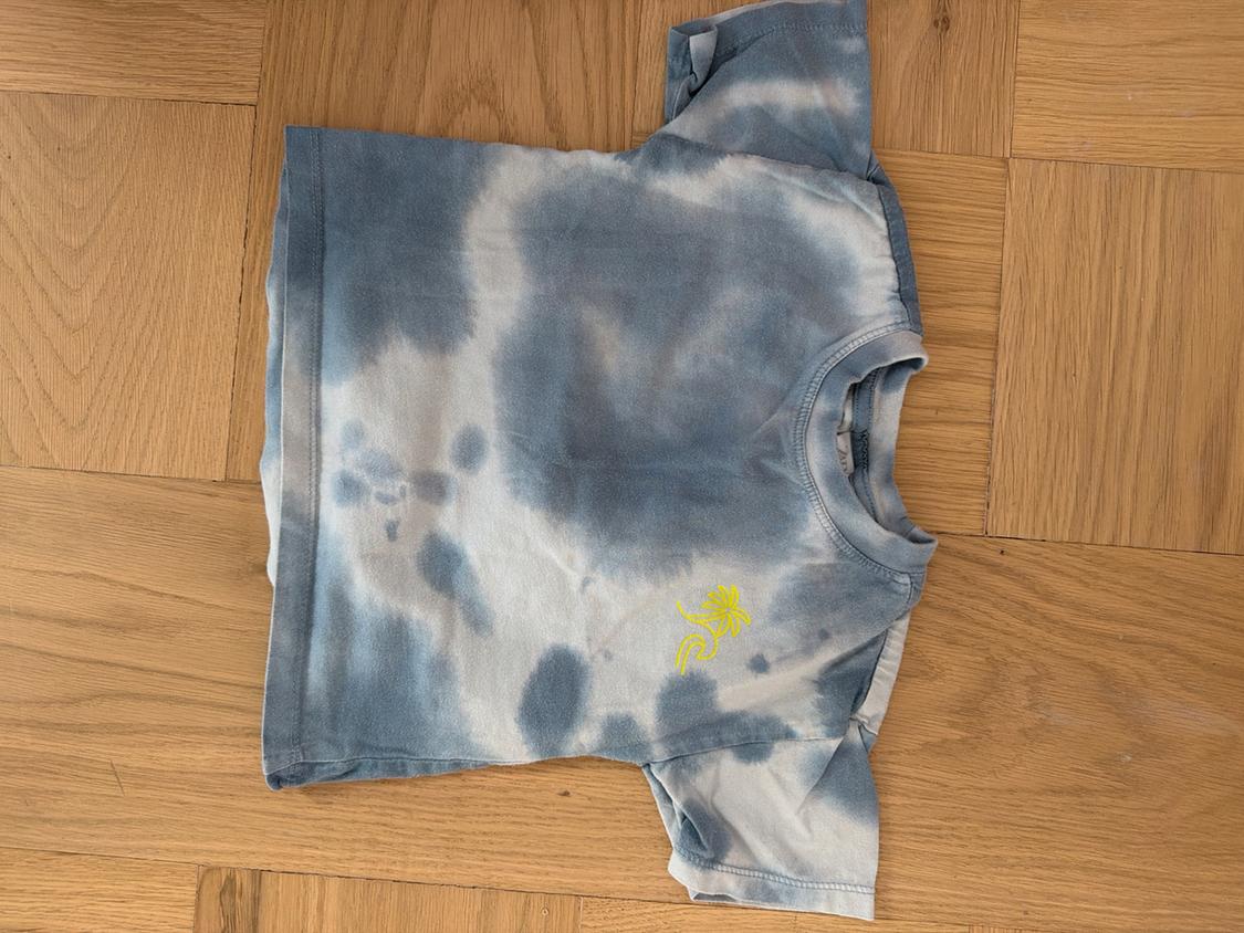 Blue and white tie-dye t-shirt, folded on a wooden floor. It has a small yellow drawing on the chest.