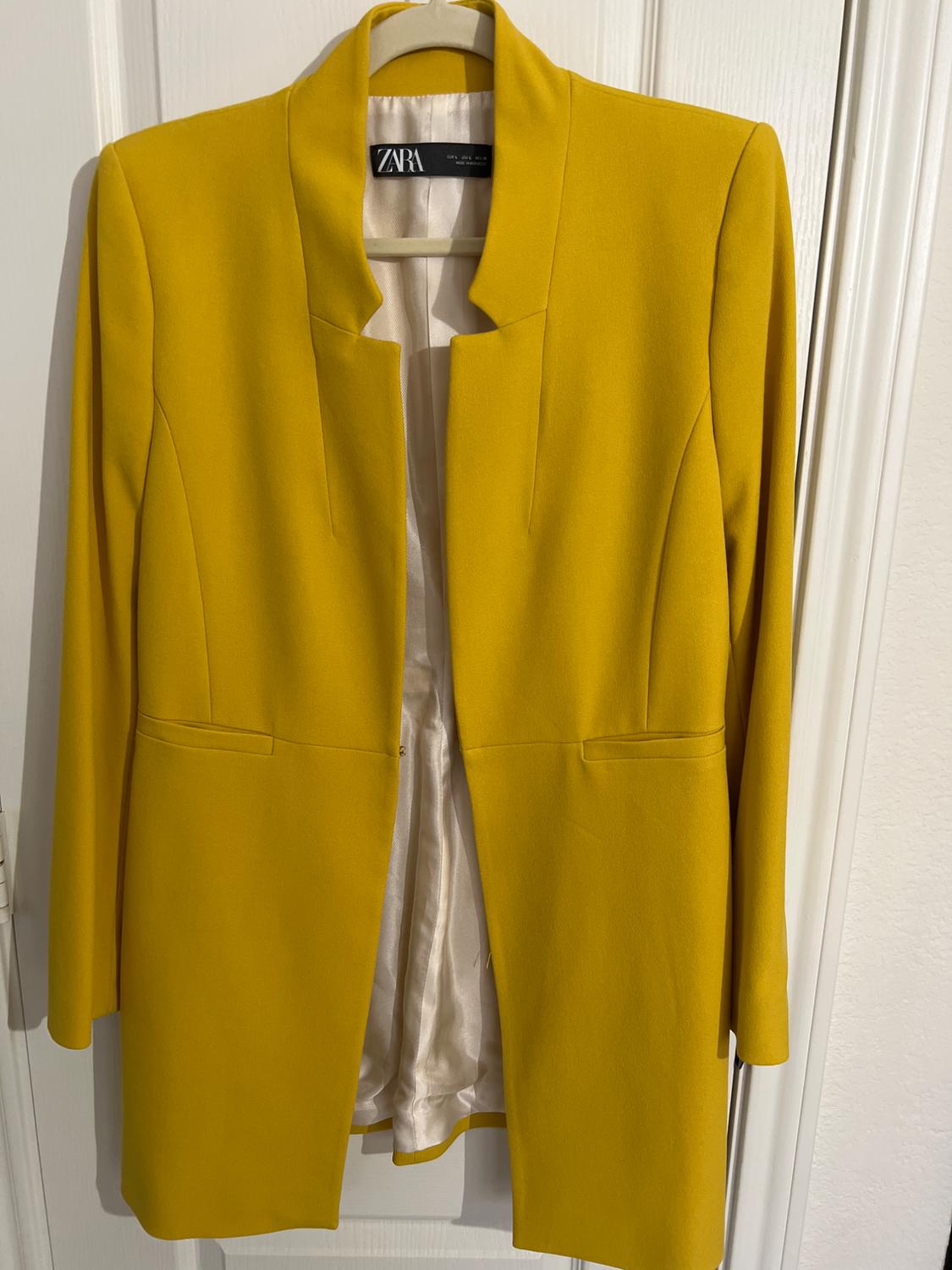 Yellow jacket hanging on a hanger, with a visible Zara tag. White door in the background.
