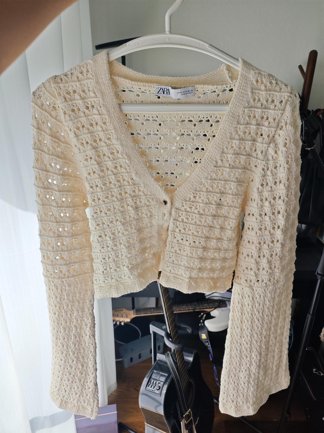 Cream open-knit cardigan hanging on a hanger. ZARA label. Room background with guitars.