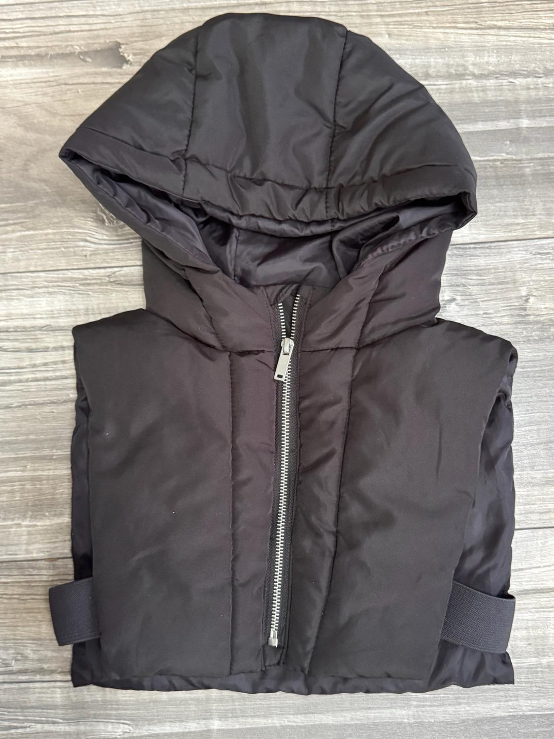 Black padded gilet with a hood, front zip and side straps, against a light wood background.