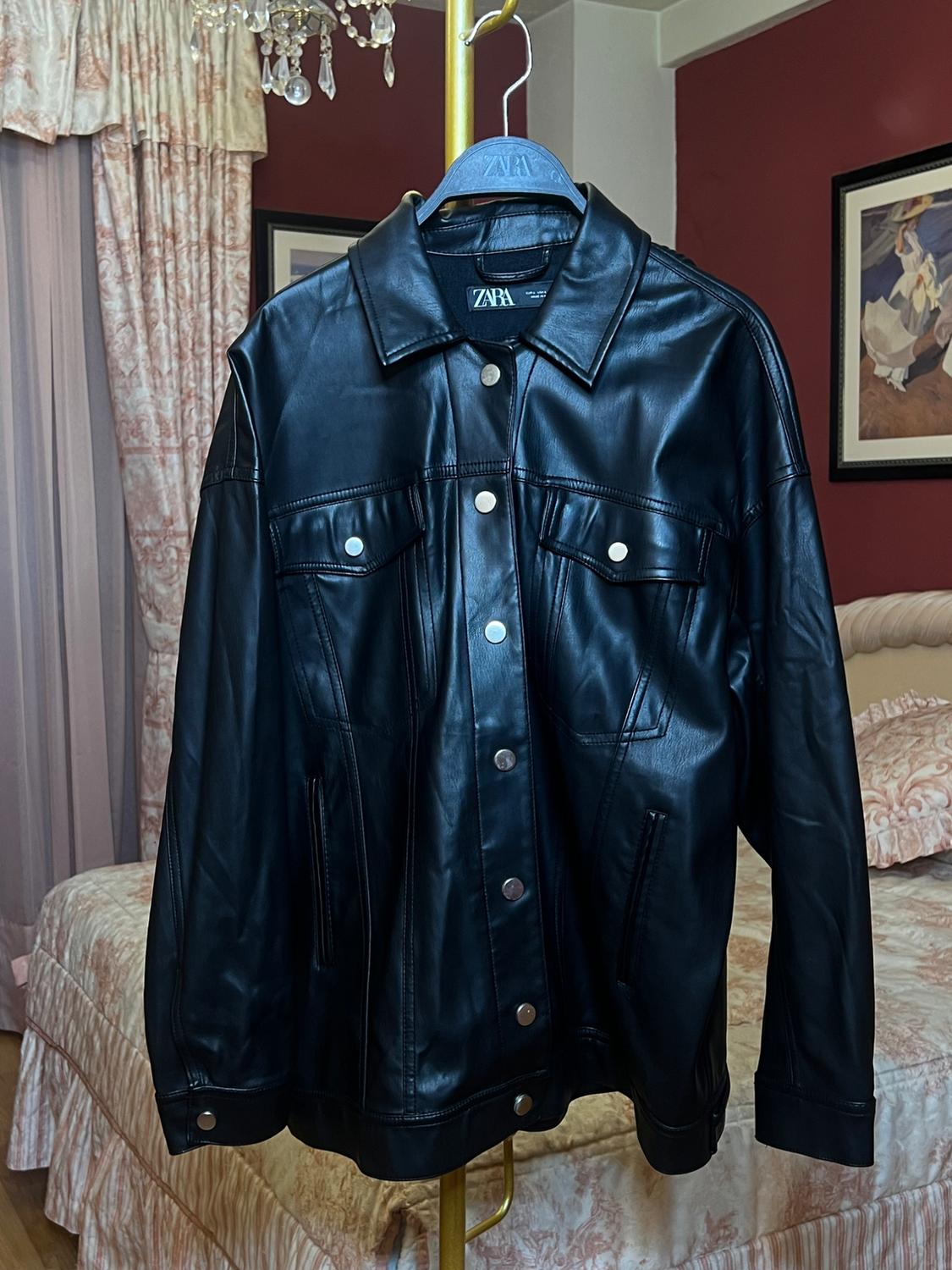 Black jacket hanging on a hanger in a bedroom with curtains and a bed with a patterned bedspread. Background with red walls and a painting.