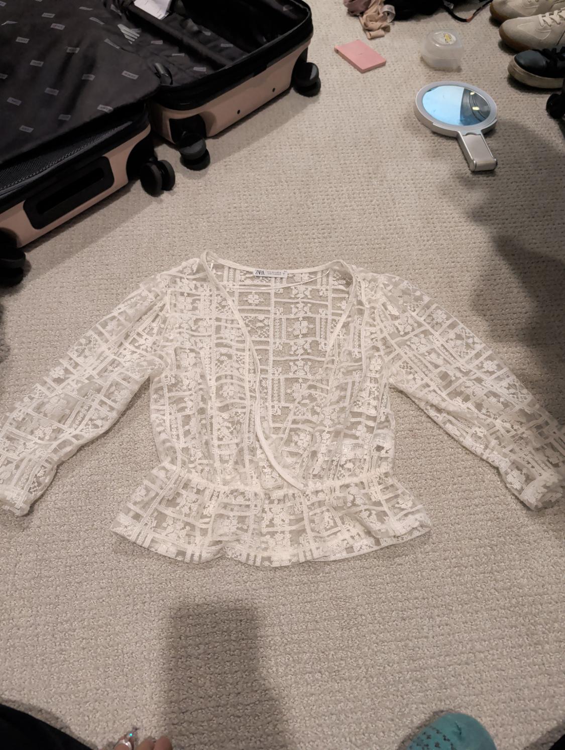 White lace blouse with long sleeves on a beige rug. Around it are an open suitcase, a hand mirror, and shoes.