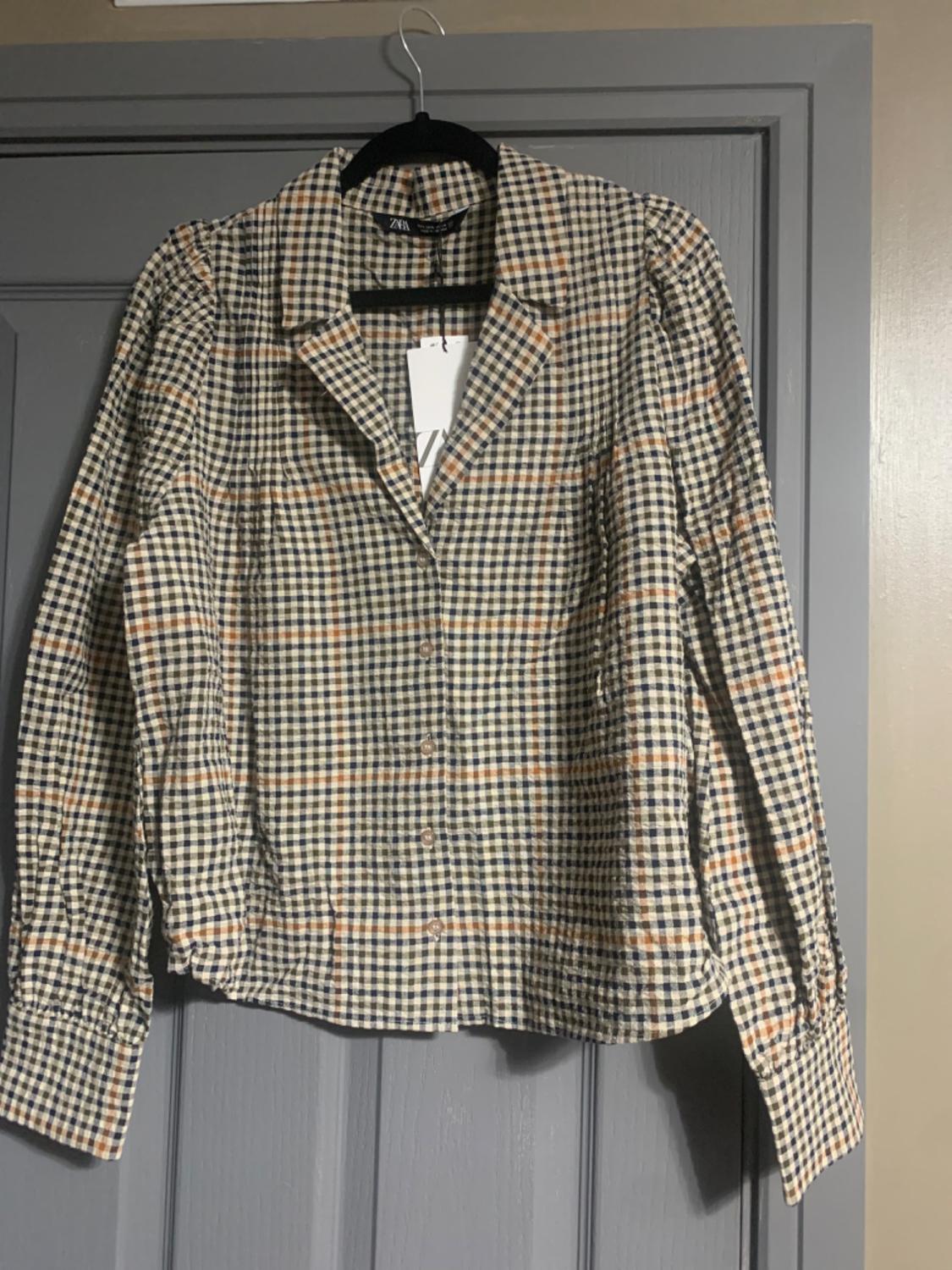 Checked shirt in shades of beige, blue and orange hanging on a black hanger over a grey door. Label visible on the collar.